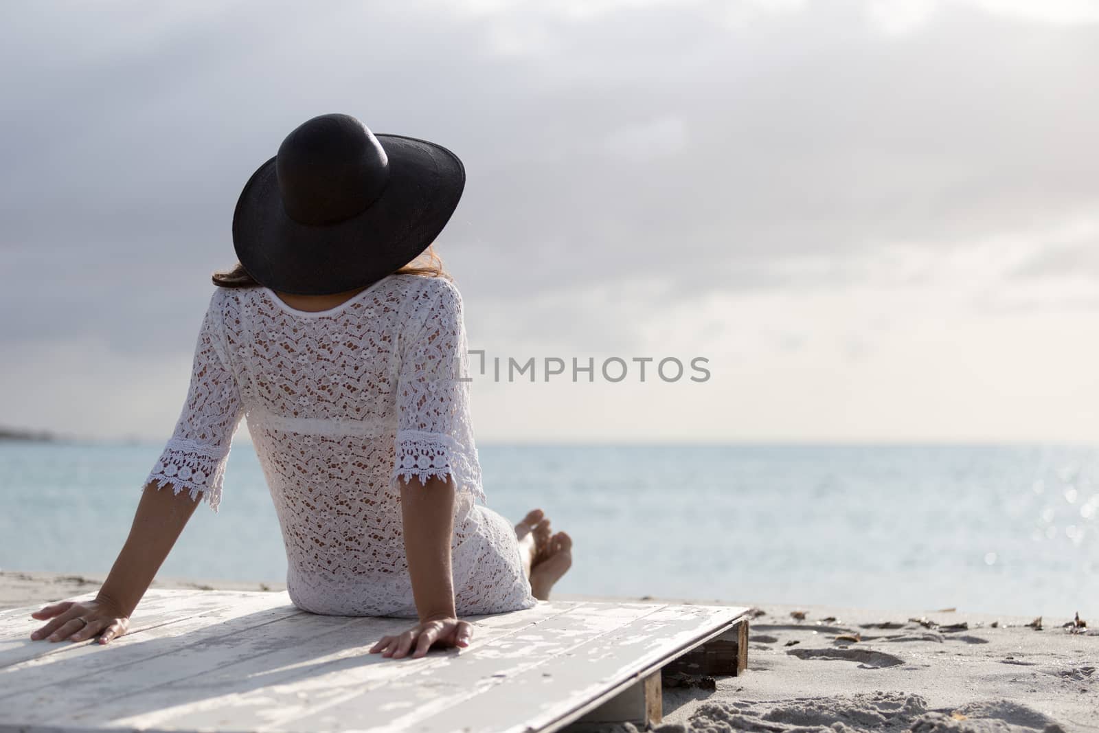 Young woman with long hair from behind sitting by the sea looks at the horizon at dawn in the wind, dressed in a white lace dress, white underwear and large black hat by robbyfontanesi