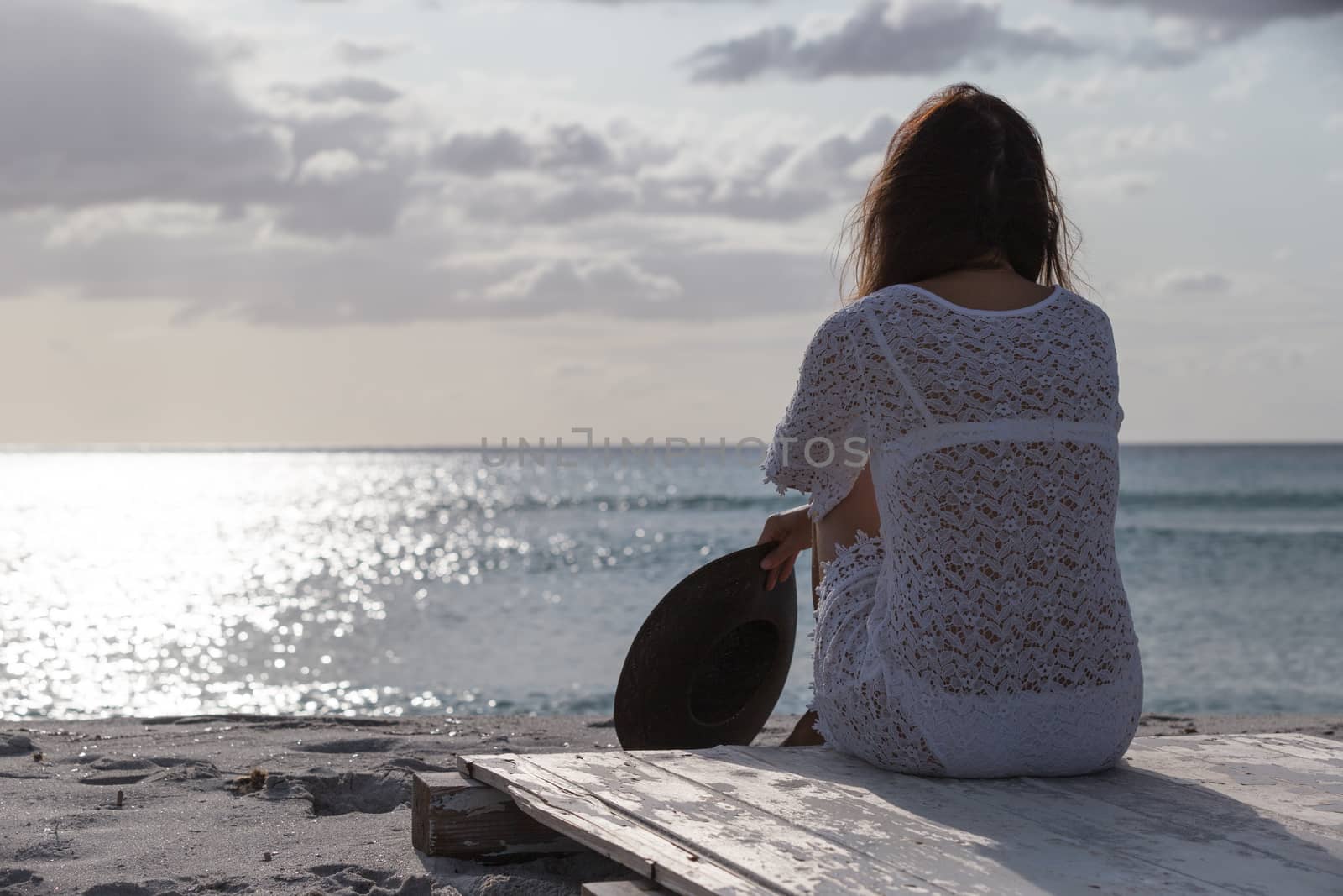 Young woman from behind sitting by the sea looks at the horizon at dawn in the wind, dressed in a white lace dress and white underwear and long hair by robbyfontanesi