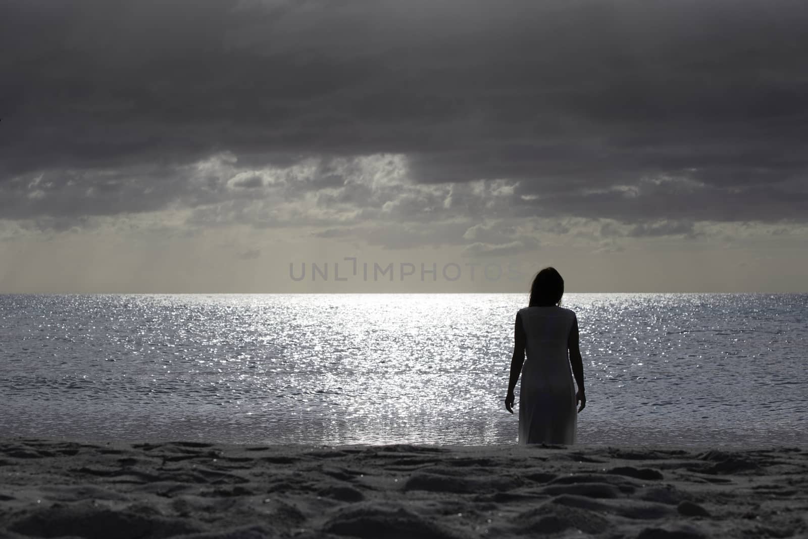 Woman silhouette from behind dressed in a light white dress walks sadly and thoughtfully towards the dark sea at dawn with dramatic sky