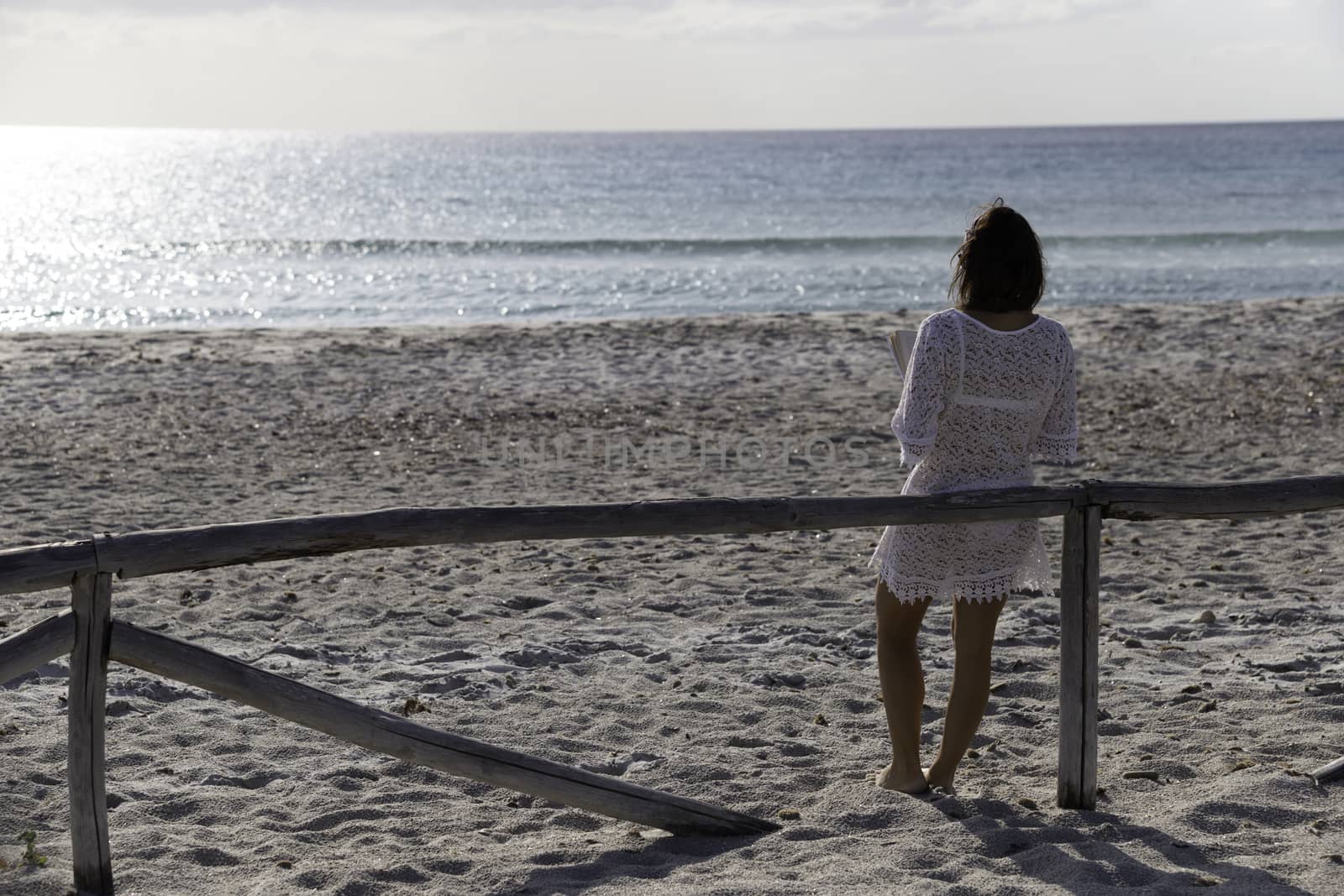 Young woman from behind reads a book on the beach looking at the horizon at dawn in the wind, dressed in a white lace dress, white underwear and long hair by robbyfontanesi