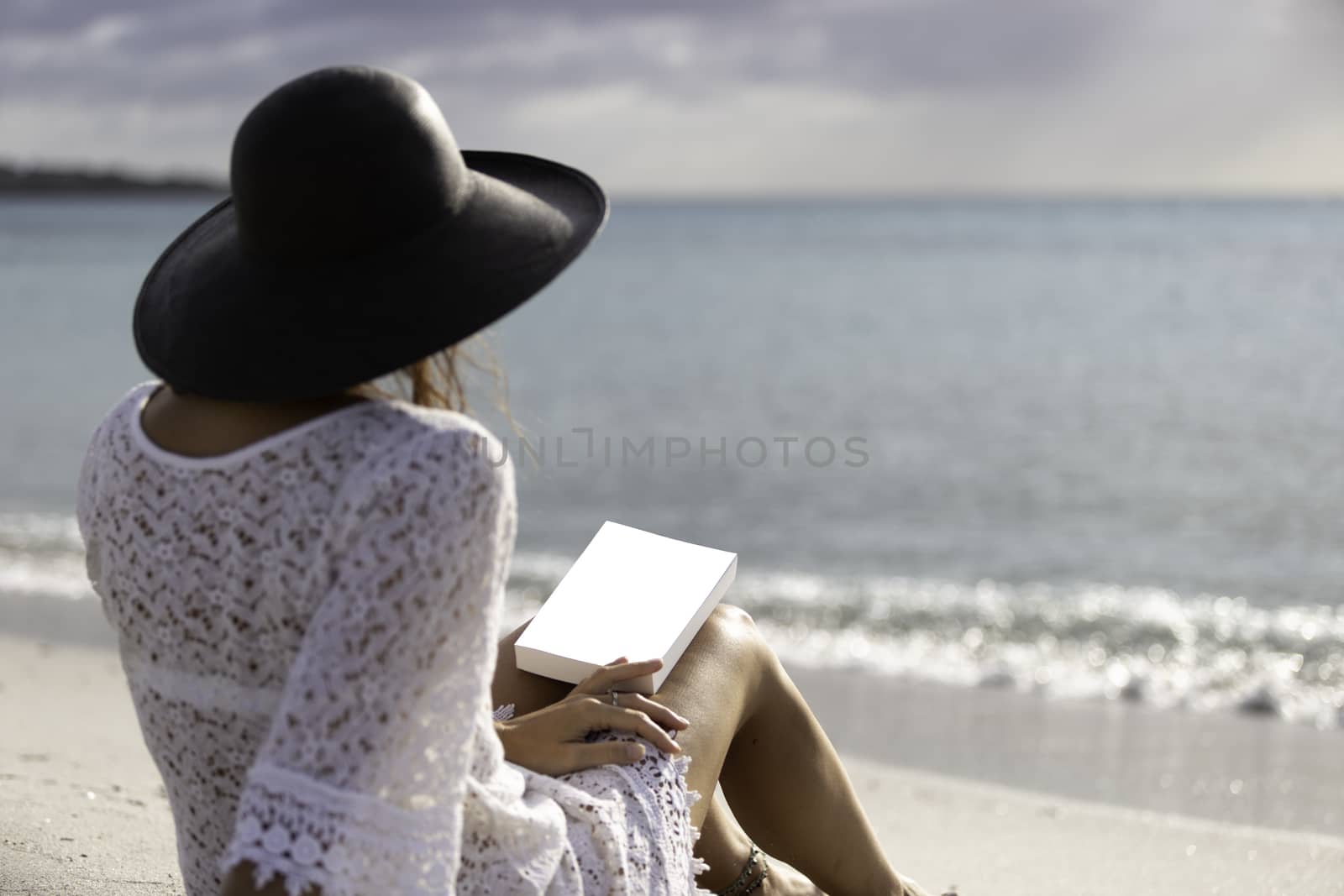 Young woman dressed in a white lace dress, white underwear and big black hat from behind sitting by the sea holding a book with blank cover on her legs by robbyfontanesi