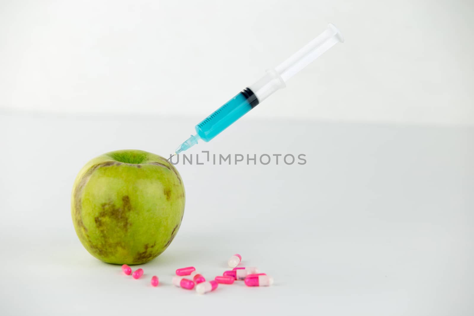 Concept: human GMO manipulation of nature and relative poisoned fruits. Close-up of an apple contaminated with a syringe threaded in and medicines on a white background