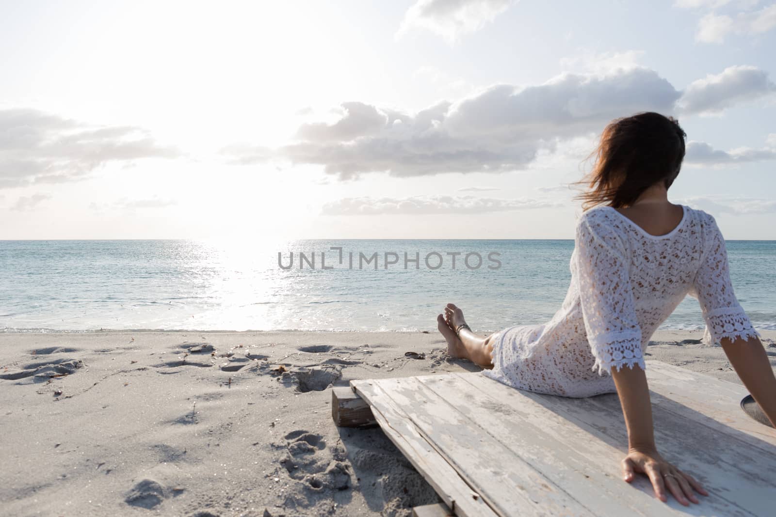 Young woman from behind sitting by the sea looks at the horizon at dawn in the wind, dressed in a white lace dress and white underwear and long hair