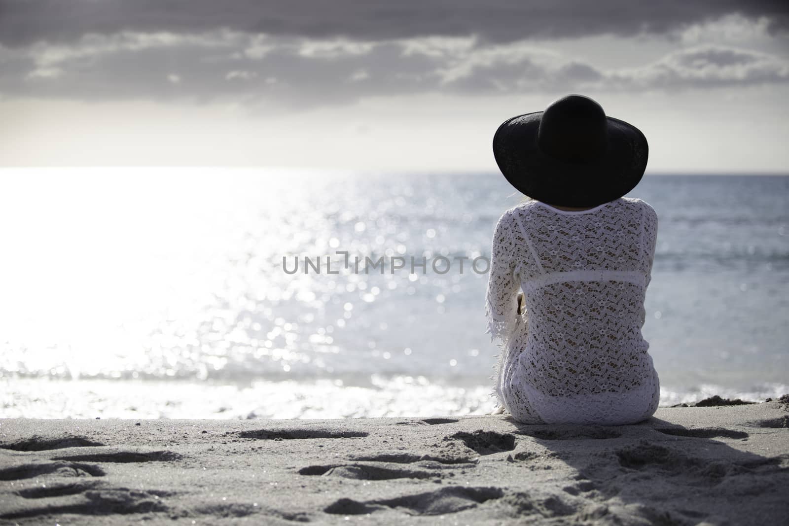 Young woman with long hair from behind sitting by the sea looks at the horizon at dawn in the wind, dressed in a white lace dress, white underwear and large black hat by robbyfontanesi