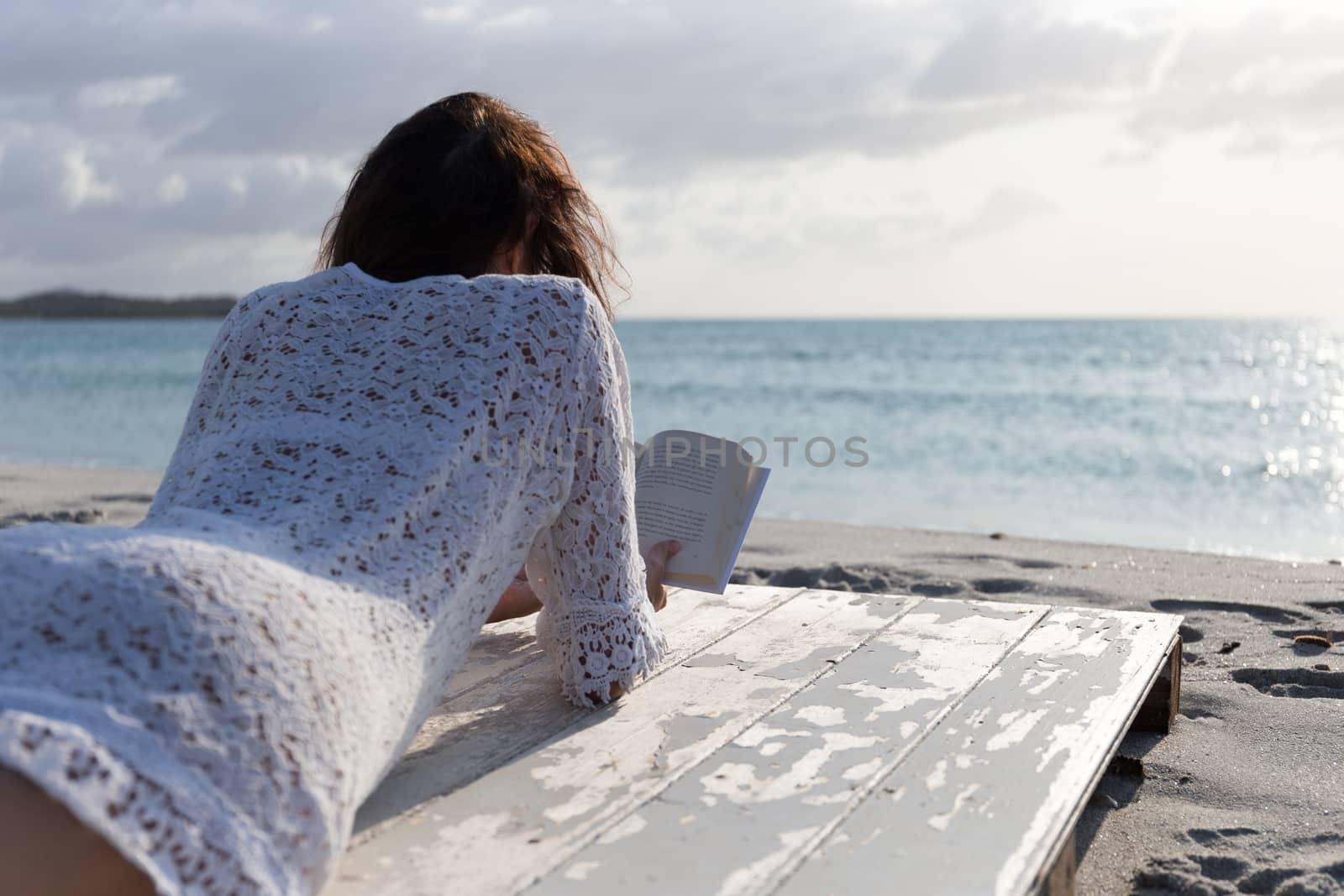 The young woman from behind lying by the sea looks at the horizon at dawn in the wind, dressed in a white lace dress and white underwear and long hair