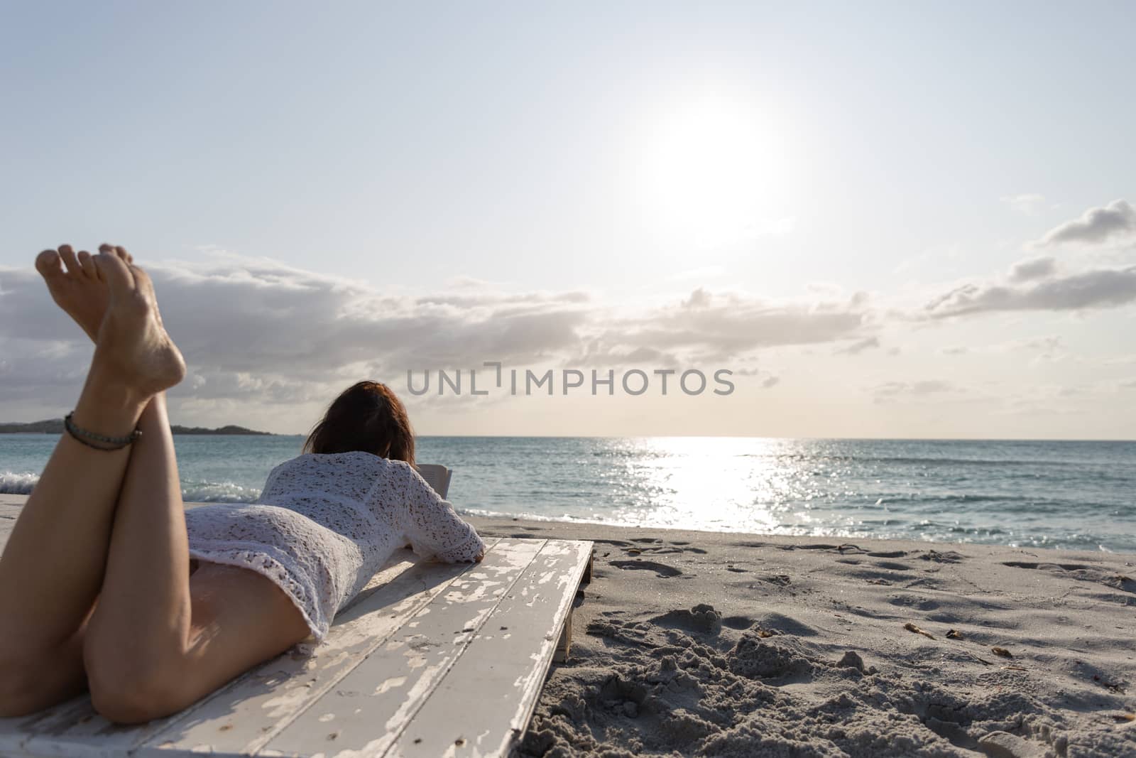 The young woman from behind lying by the sea looks at the horizon at dawn in the wind, dressed in a white lace dress and white underwear and long hair