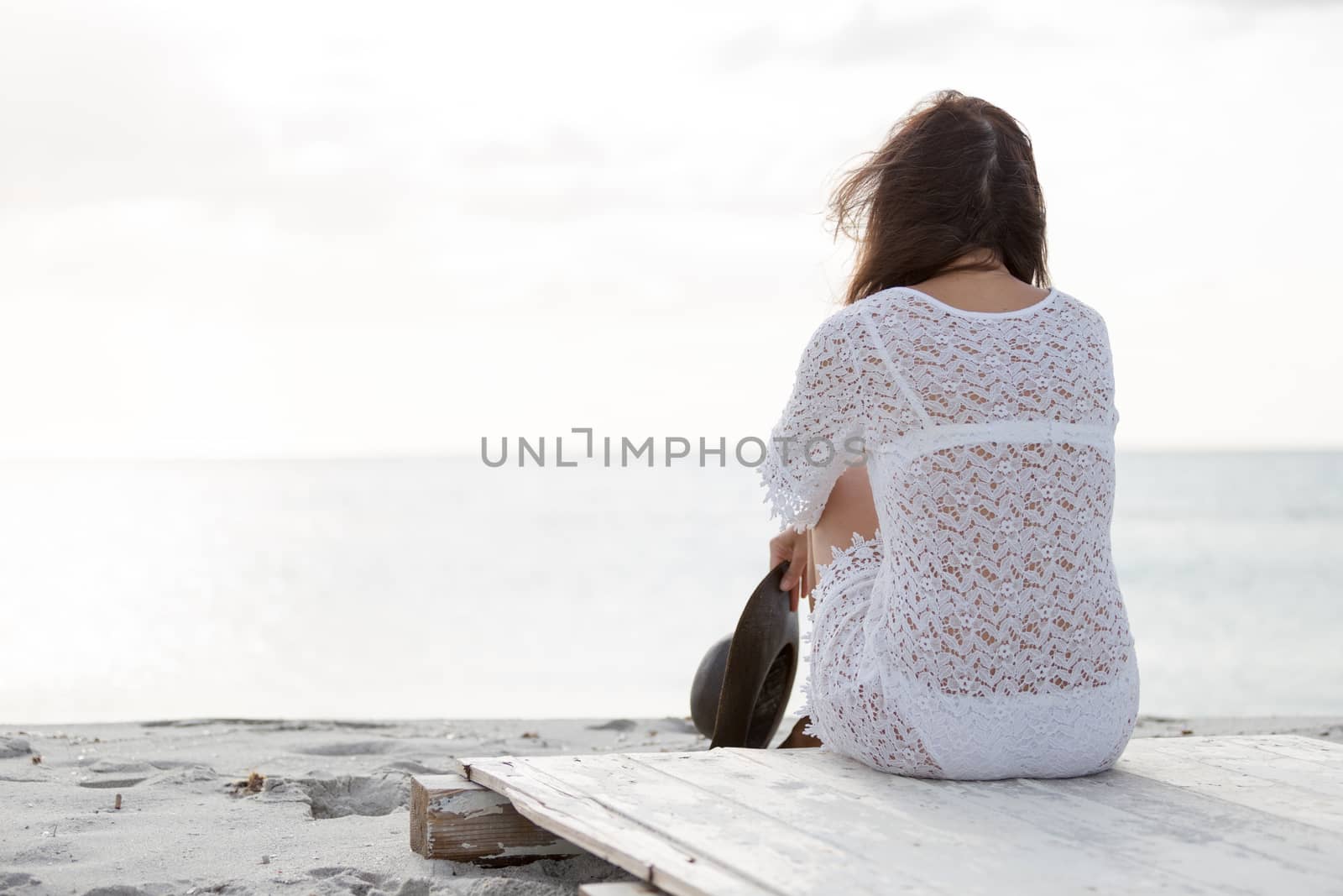 Young woman from behind sitting by the sea looks at the horizon at dawn in the wind, dressed in a white lace dress and white underwear and long hair