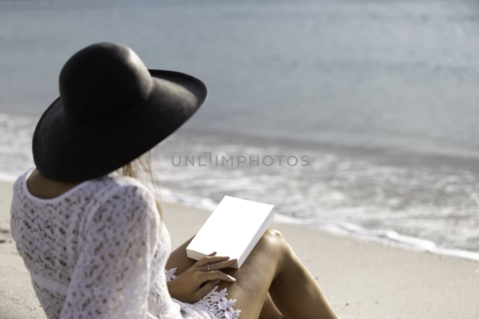 Young woman dressed in a white lace dress, white underwear and big black hat from behind sitting by the sea holding a book with blank cover on her legs by robbyfontanesi