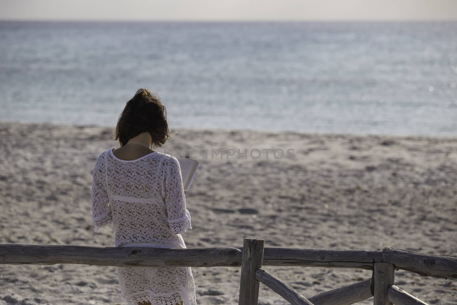 Young woman from behind reads a book on the beach looking at the horizon at dawn in the wind, dressed in a white lace dress, white underwear and long hair by robbyfontanesi