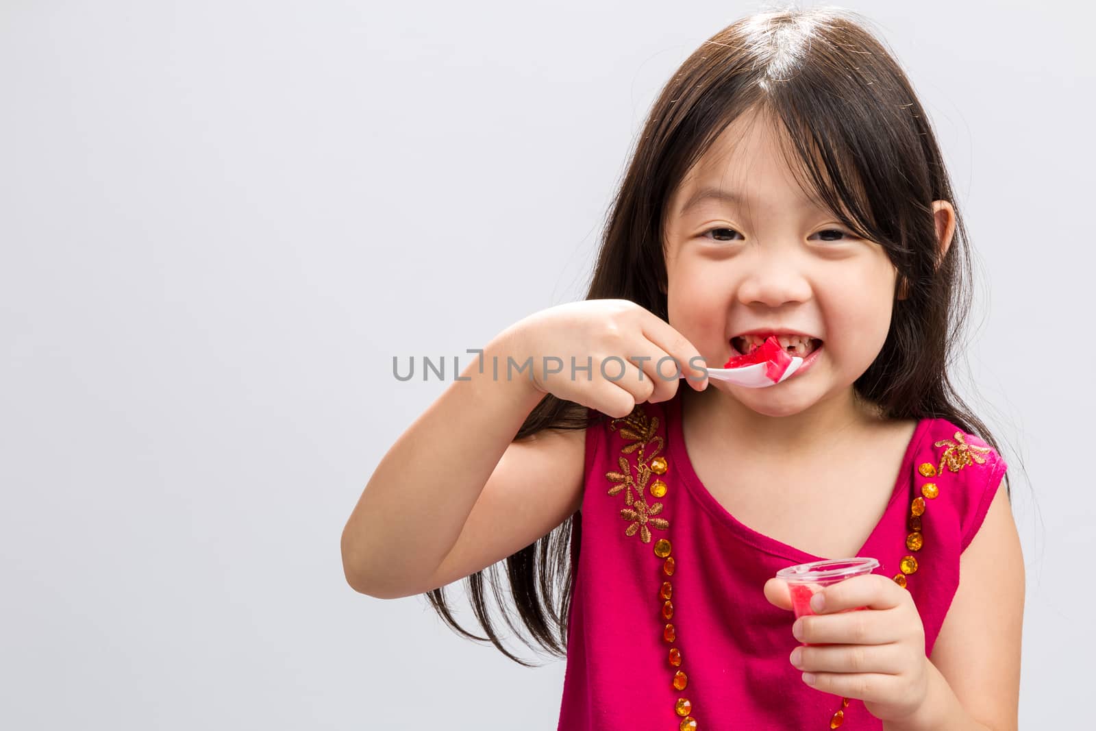 Young girl happily eating cup of gelatin dessert on isolated white background.