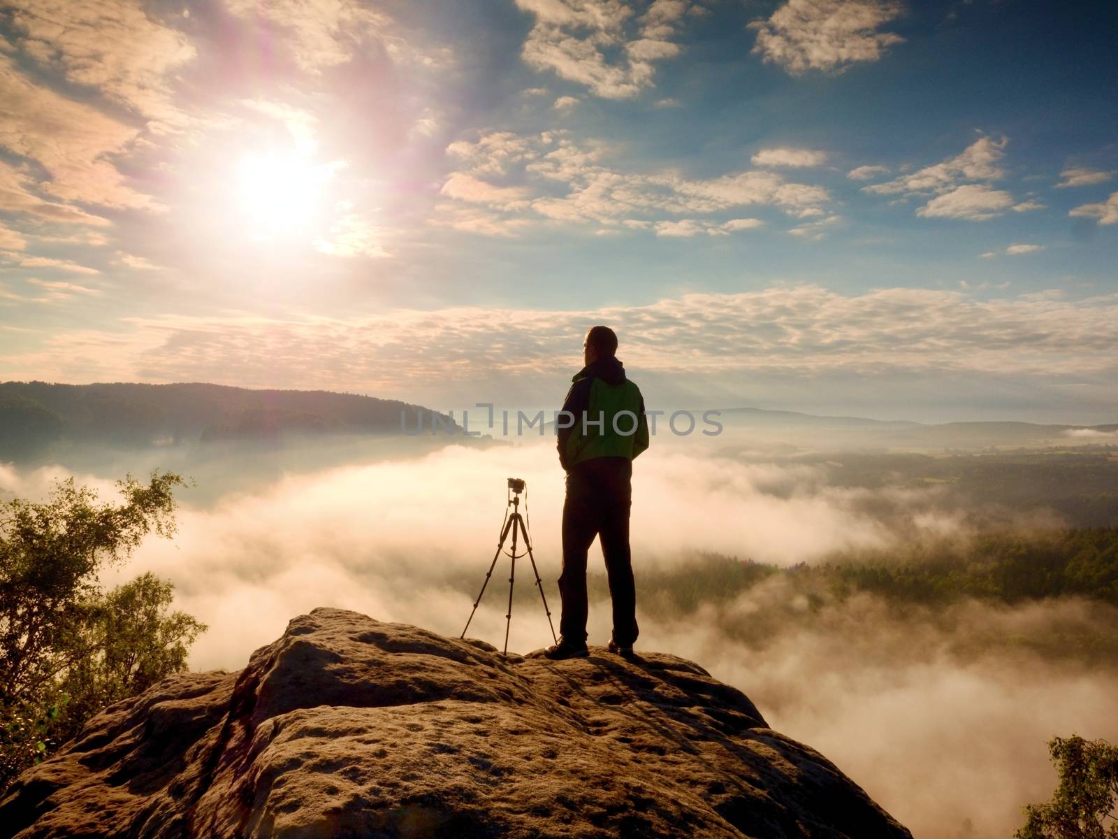 Professional photographer with tripod on cliff and thinking. Dreamy fogy landscape, blue misty sunrise in a beautiful valley below