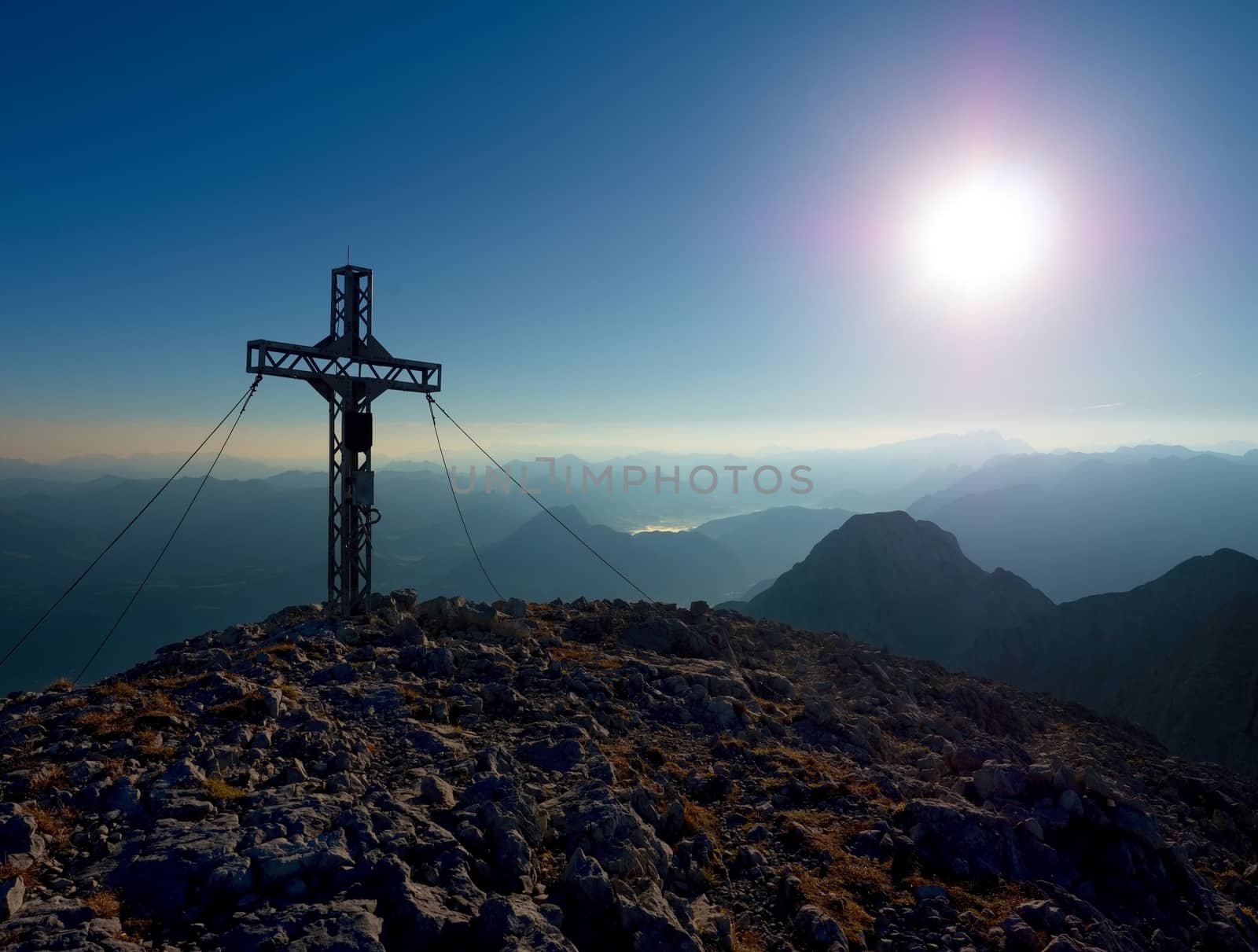 Steel crucifix at mountain peak in Alps. Sharp rocky summit, daybreak Sun in sky.  by rdonar2