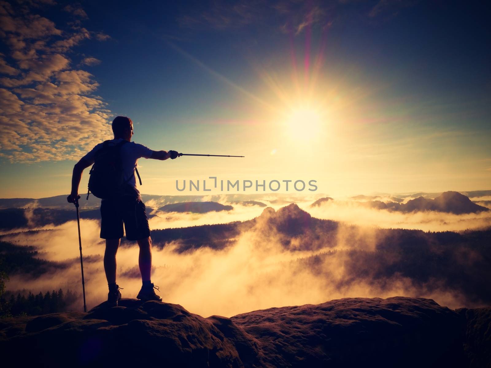 Silhouette of tourist with poles in hand. Sunny spring landscape,  rocky summit. Hiker with sporty backpack stand on rocky view point above misty valley. 