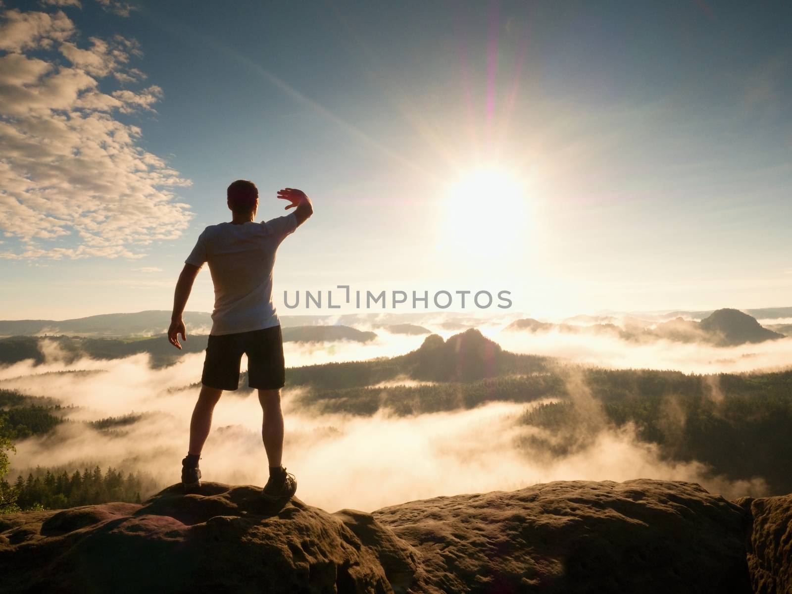 Young sportsman shadowing his eyes from the bright light of the daybreak Sun. Mountain summit achievement. Colorful misty morning in rocks.