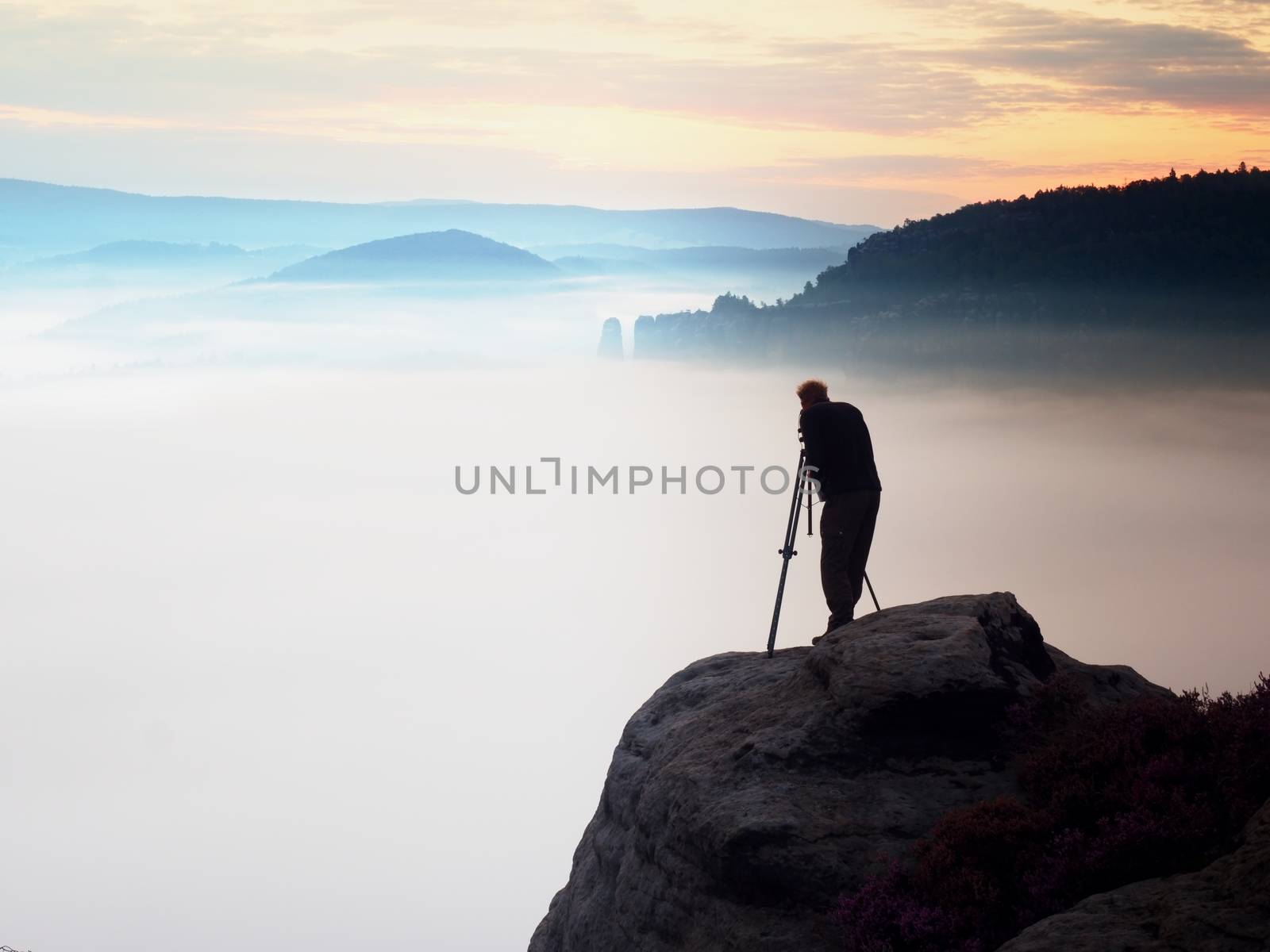 Professional photographer takes photos with mirror camera and tripod on peak of rock. Dreamy fogy landscape, spring orange pink misty sunrise in beautiful valley below.