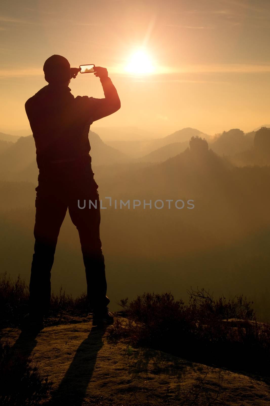 Man takes photos with phone on rock empire. Dreamy fogy mountains, spring orange pink misty sunrise in a beautiful valley of rocky mountains.