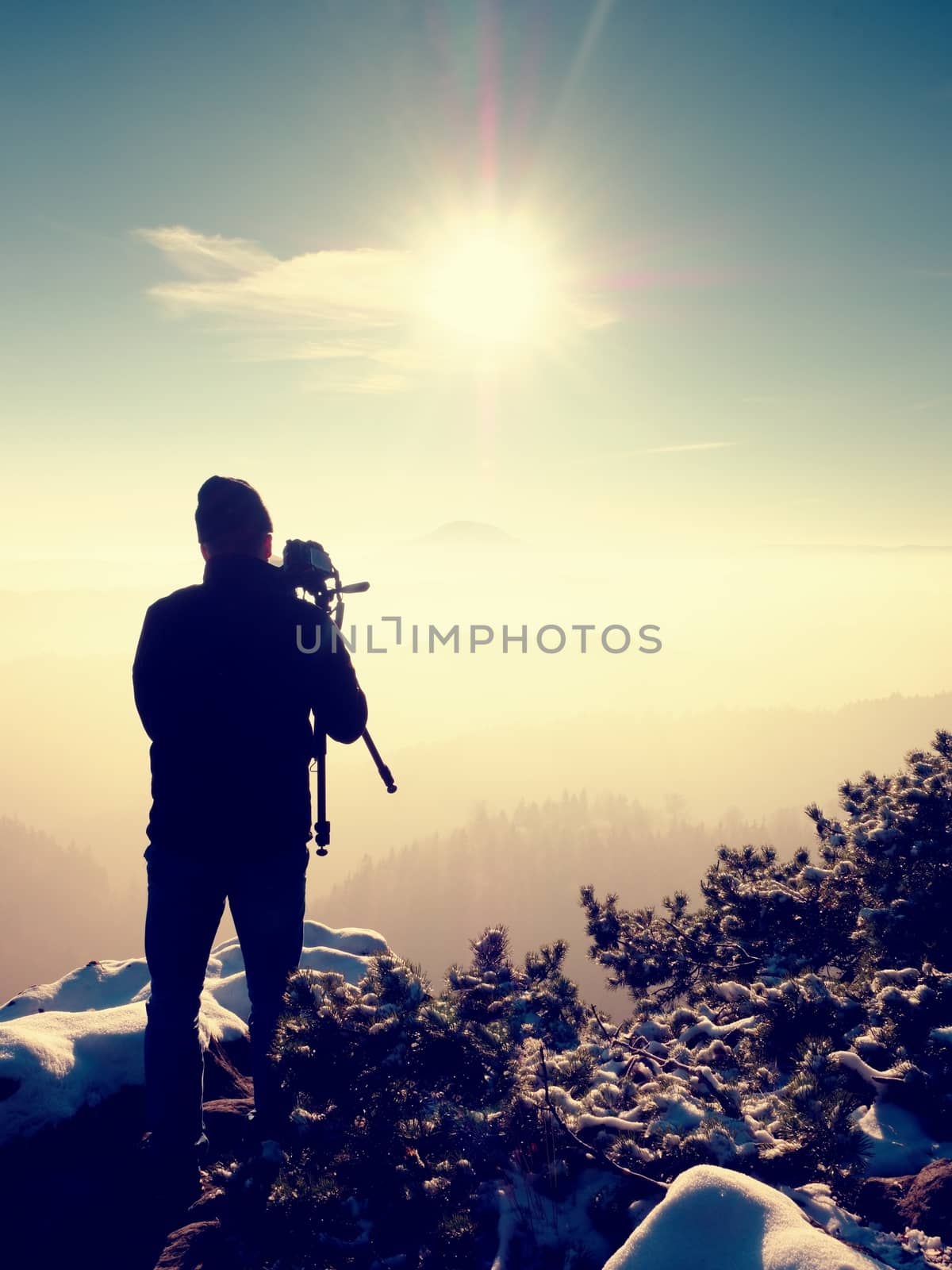 Professional nature photographer do work on snowy cliff.  Man takes photos with mirror camera on peak of rock. Dreamy fogy landscape, spring orange pink misty sunrise.