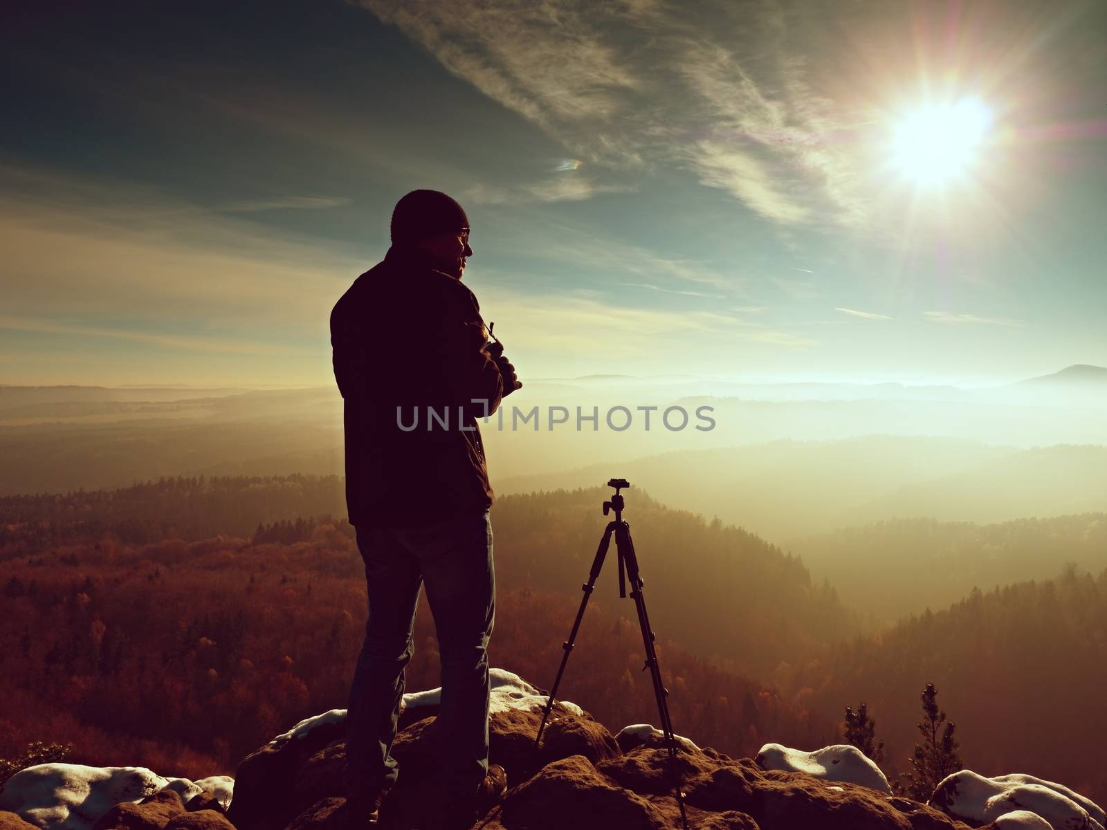 Nature photographer stay with camera ind hands  at tripod on rock and thinking. First snow. Dreamy fogy landscape, orange misty sunrise in  beautiful mountains