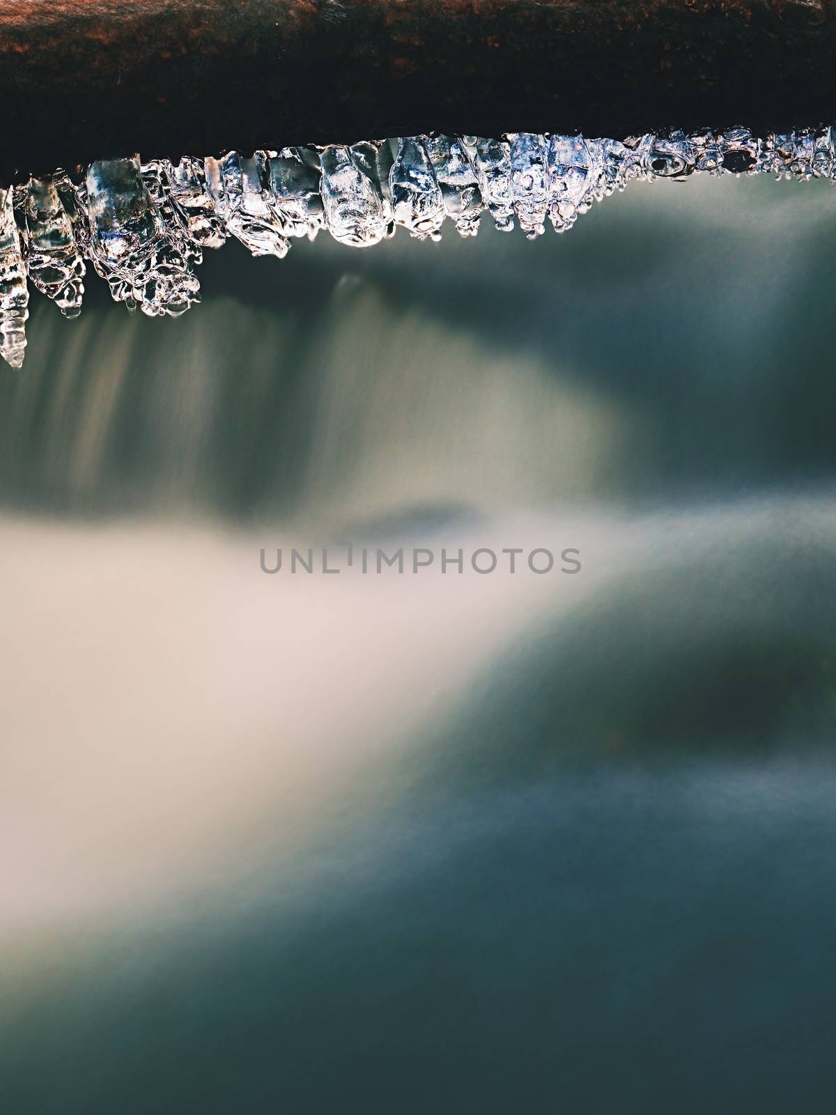 Long icicles hang above dark freeze  water of mountain stream. Winter season at river, thin icicles are hanging on fallen trunk above milky water level. 