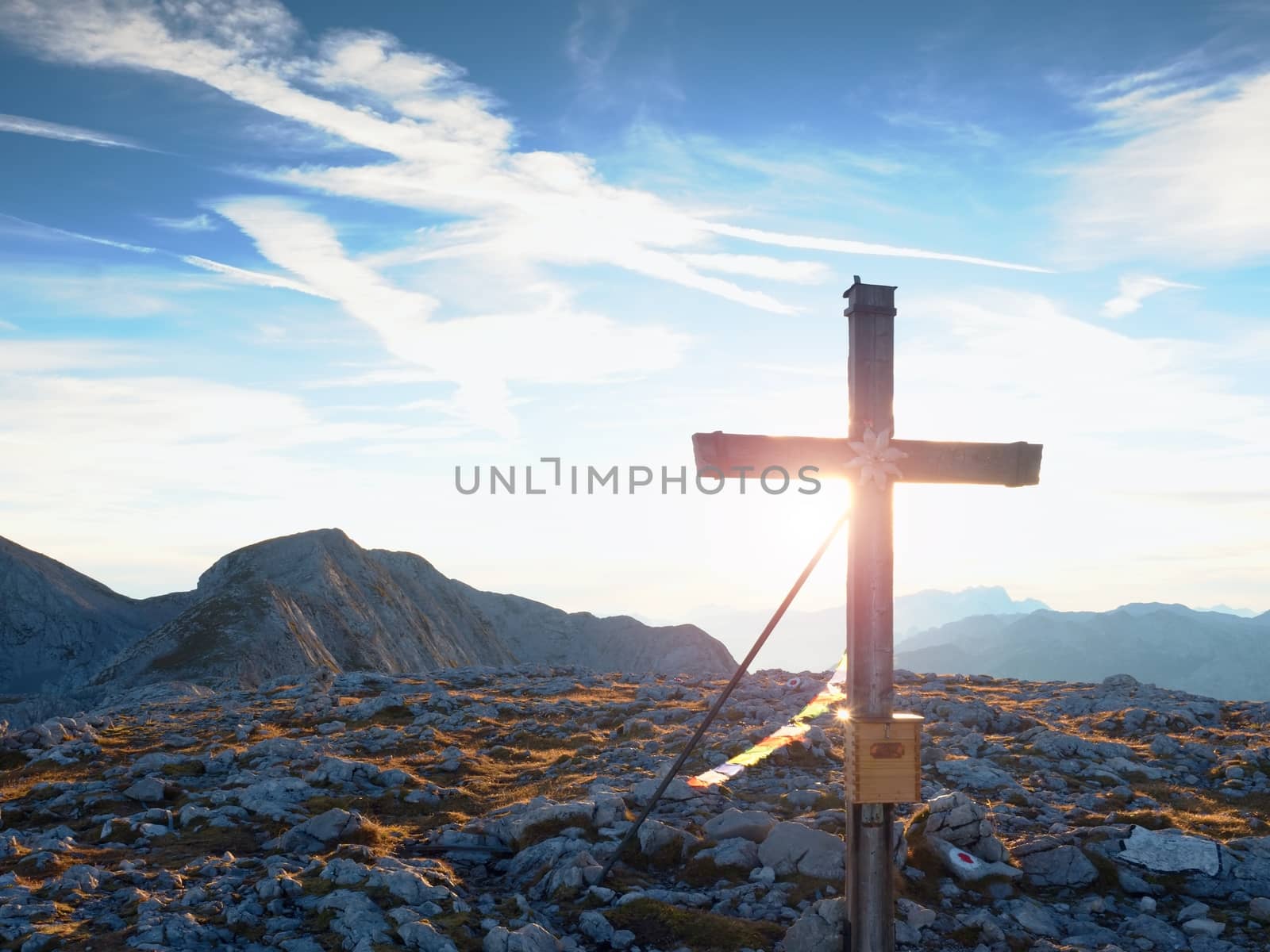 Modest wooden cross  with Buddhist praying flags, raised on rocky Alpine mountain summit . Sharp rocky peak. Gentle clouds  in blue sky. Wooden unpretentious crucifix in memory of victims of mountains.