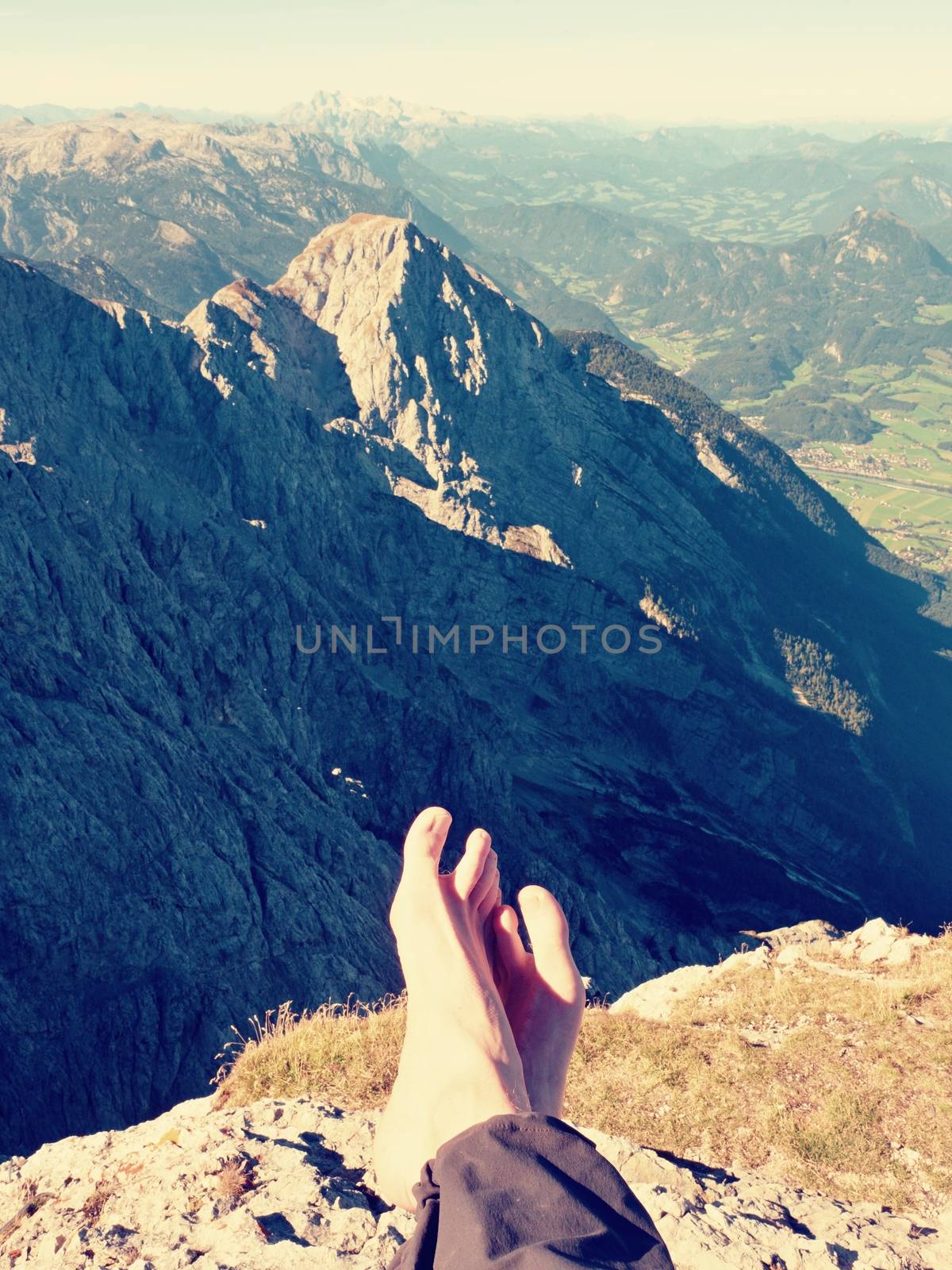 Naked male sweaty legs in dark hiking trousers take a rest on peak of mountain above spring valley. by rdonar2