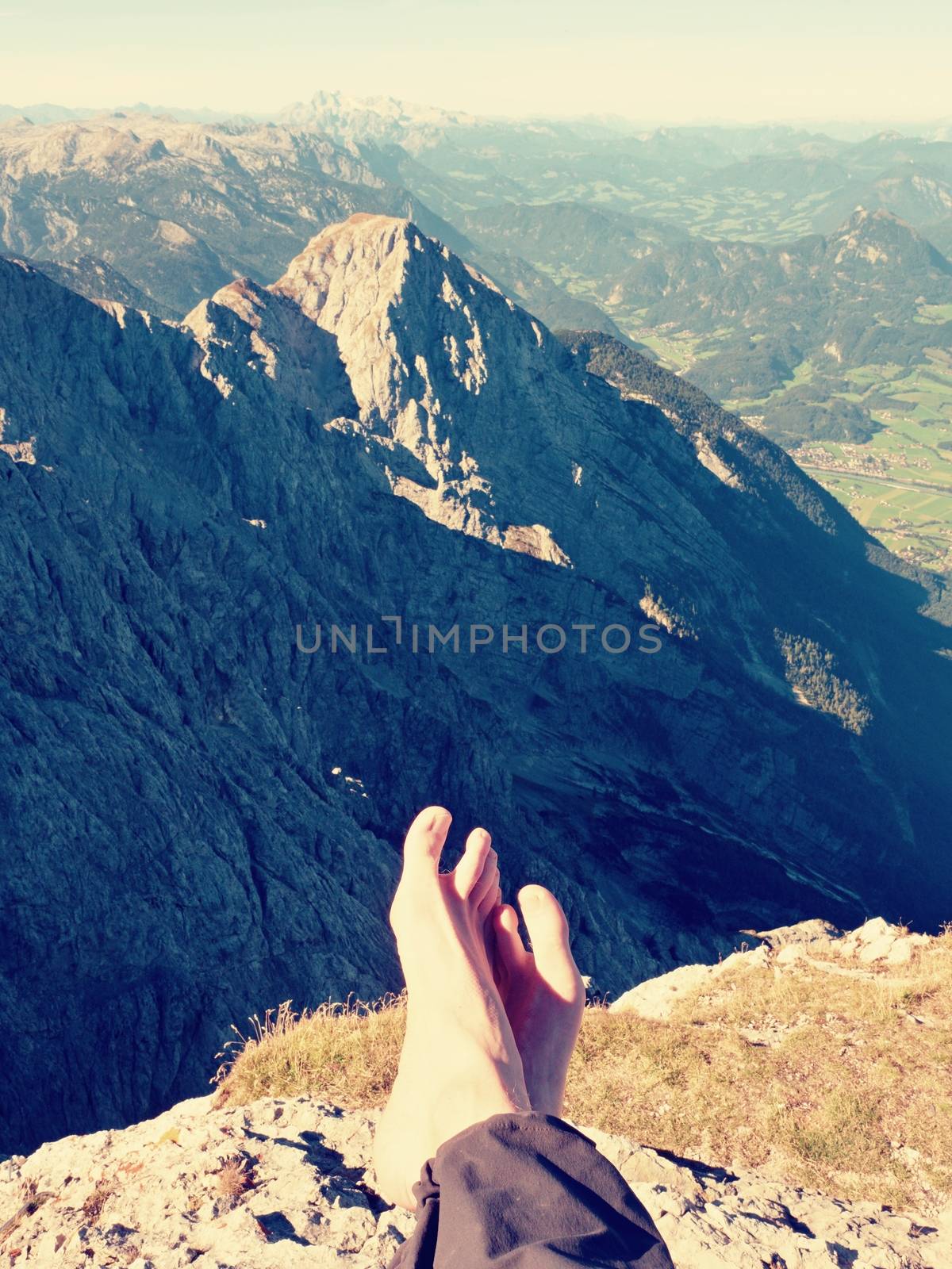 Naked male sweaty legs in dark hiking trousers take a rest on peak of mountain above spring valley. The rest on tiring mountain trail.