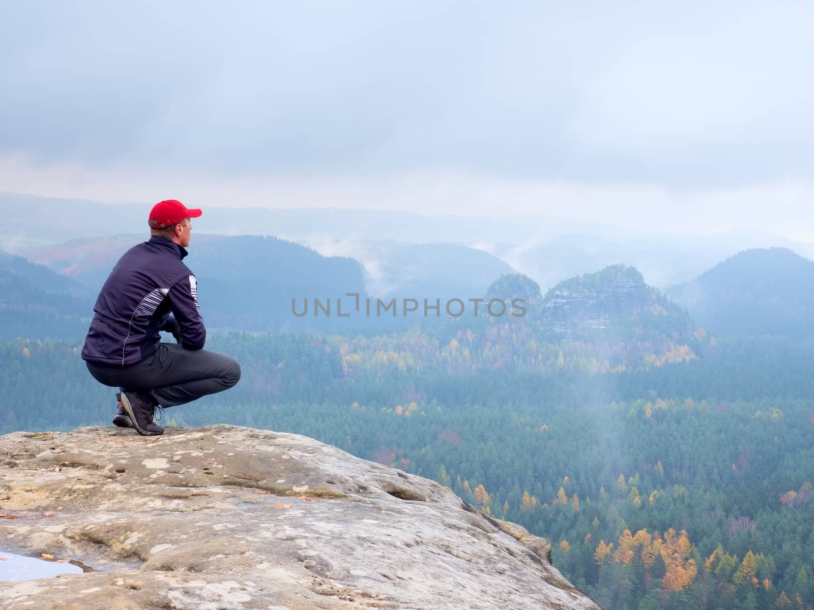 Hiker in red cap and  black sportswear in squatting position on a rock, enjoy autumn scenery. Long valley full of heavy creamy fog after rainy night