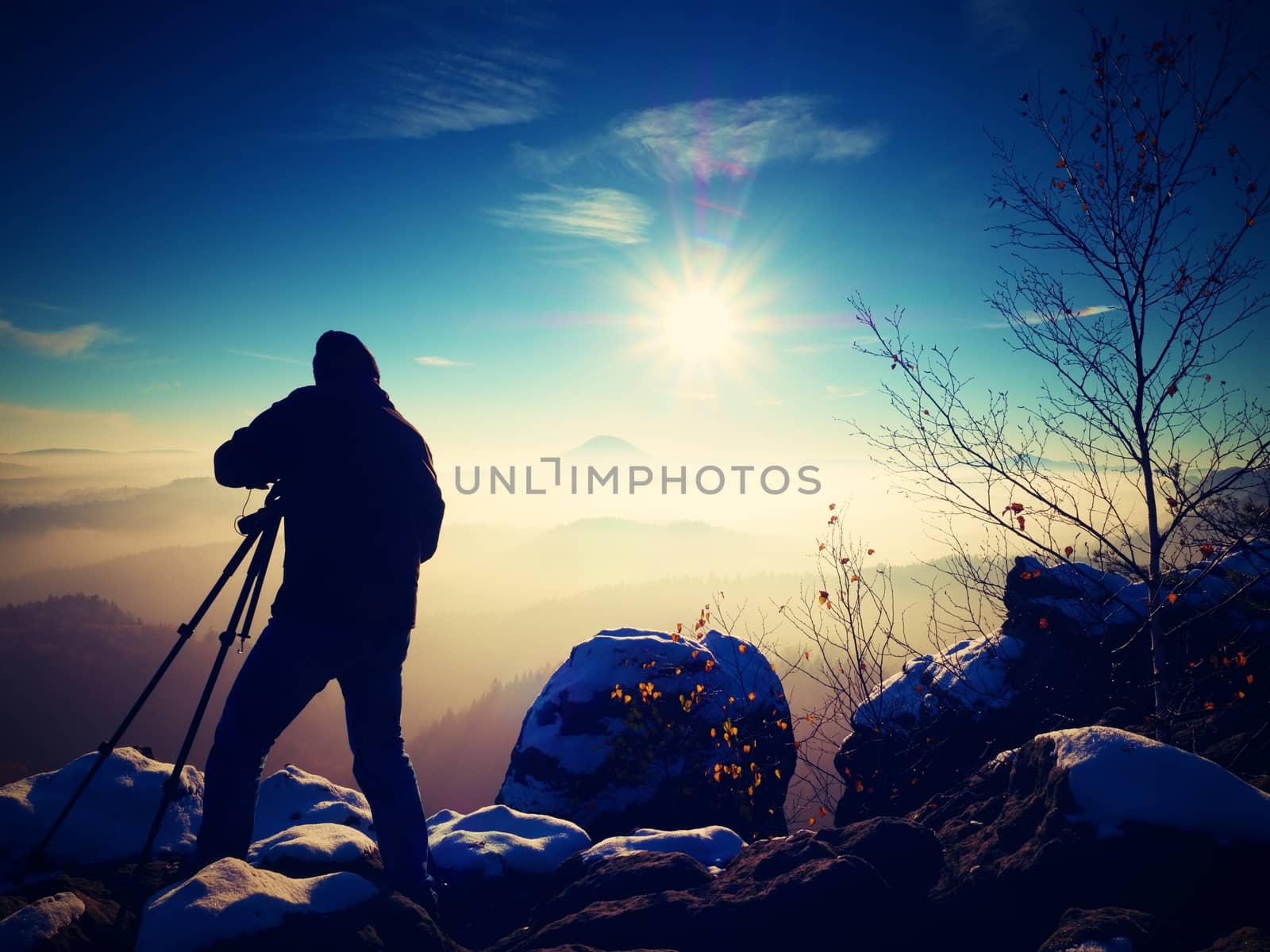 Sunny early winter  morning. Photographer preparing camera on tripod. Snowy rocks, in valley bellow colorful leaves forest. View over misty and foggy valley to Sun.