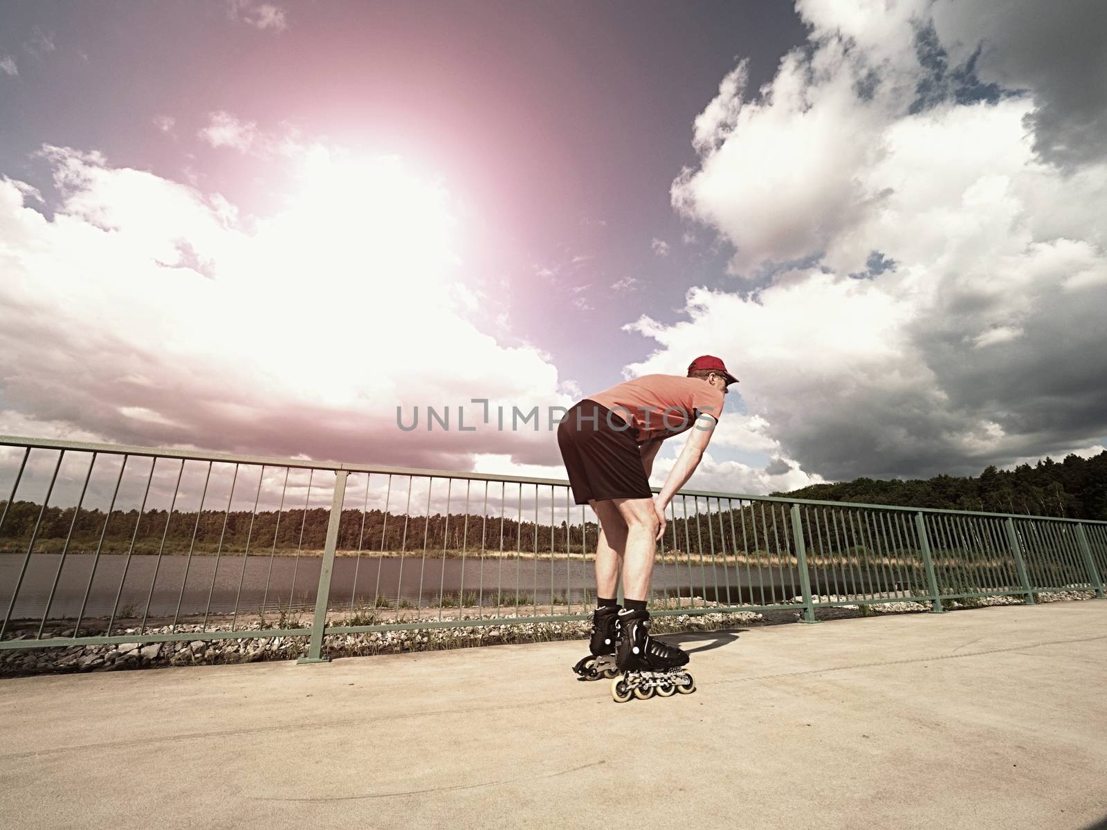 Middle age man in red t-shirt  with inline skates ride in summer park, popular outdoor roller skating. Sea bridge with blue sky in backgrround