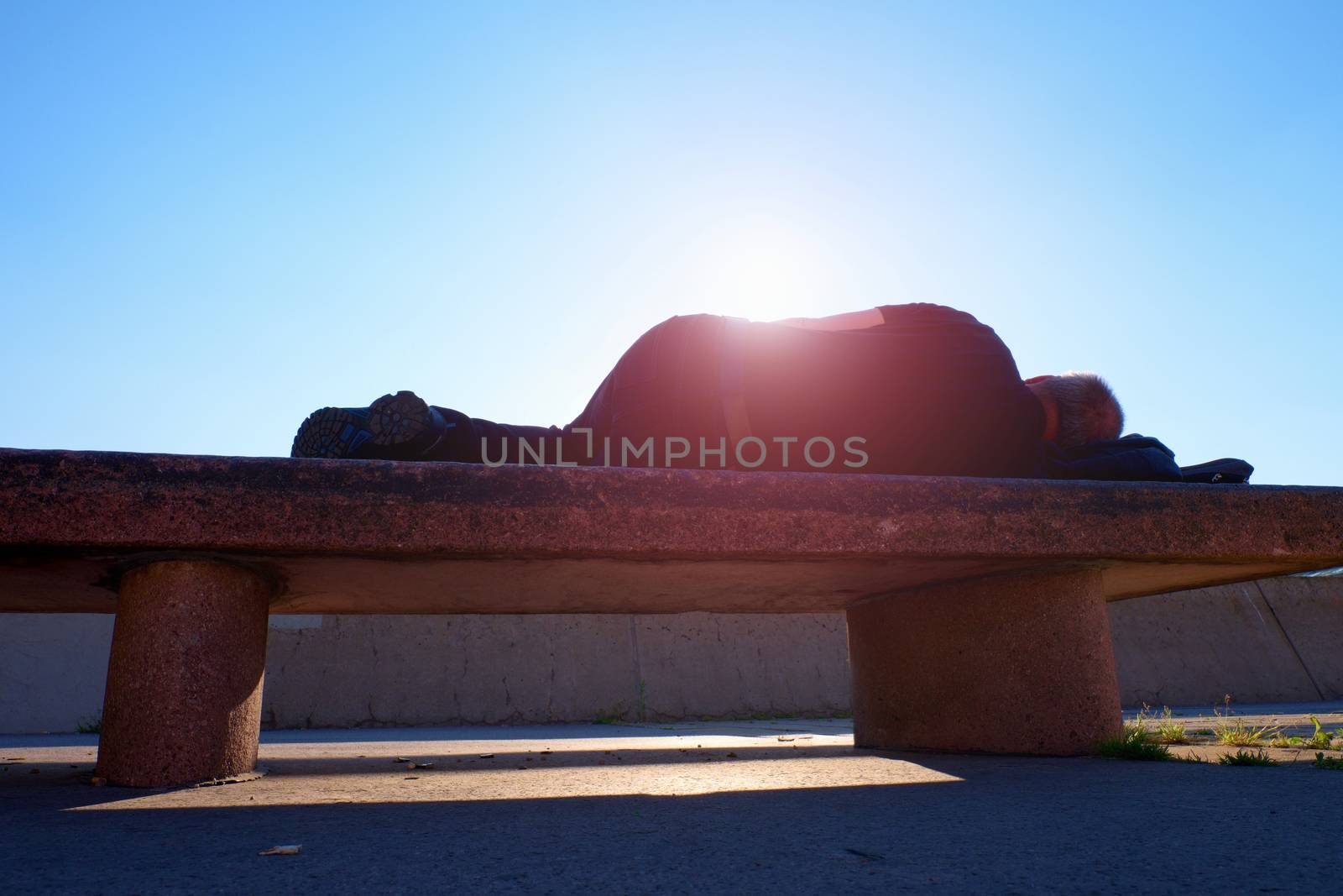 Drank adult man rests on stony  bench in park, Sharp sun rays make contours around sleeping body. 
