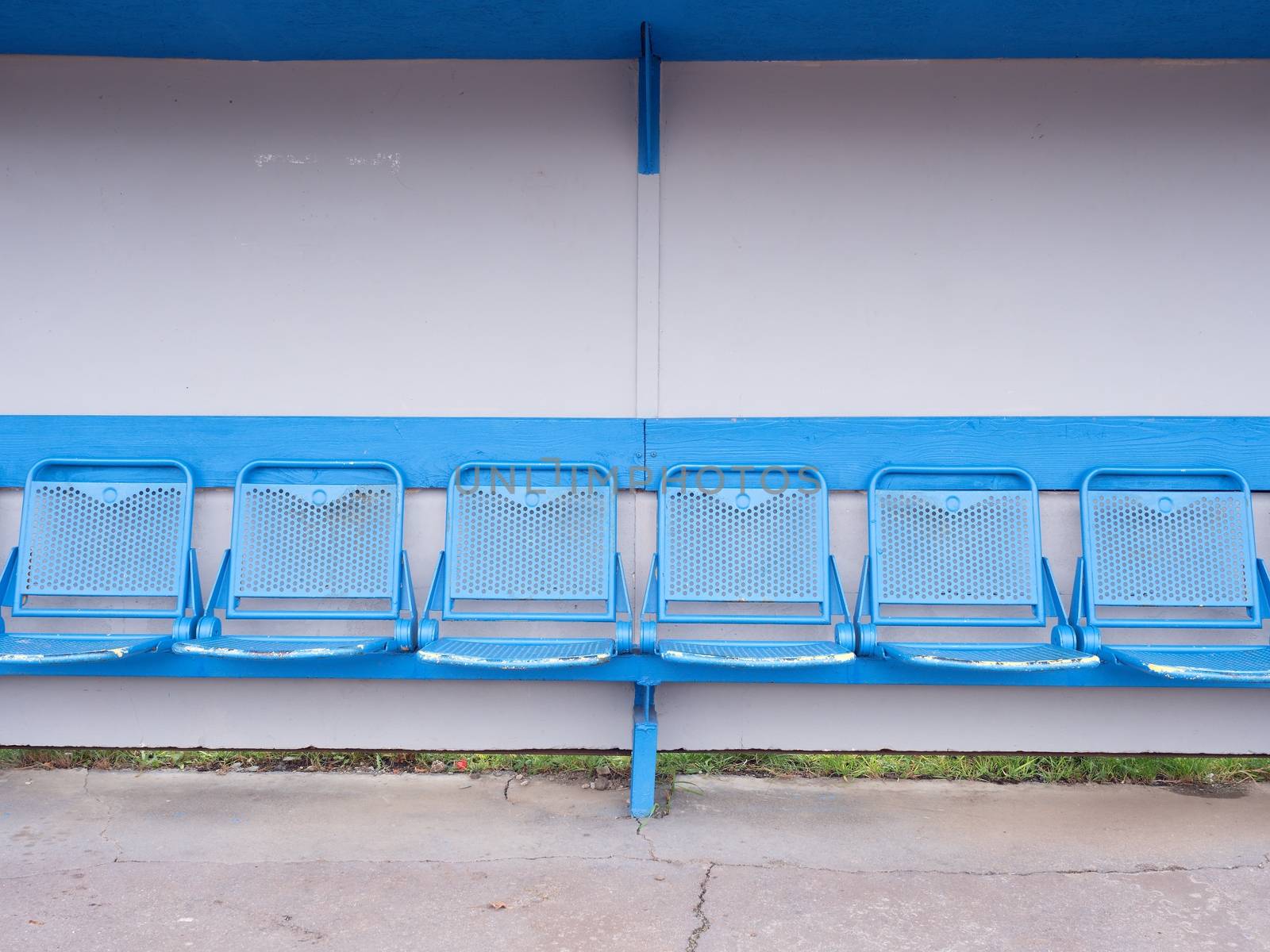 New blue metal seats on outdoor stadium players bench, chairs with blue paint below wooden roof. 
