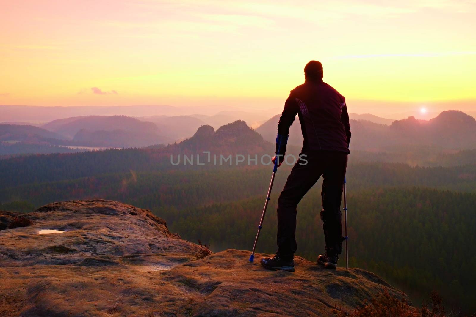 Hiker with broken leg in immobilizer. Tourist with  medicine crutch on mountain peak. Deep misty valley bellow silhouette of man with hand in air. Spring daybreak