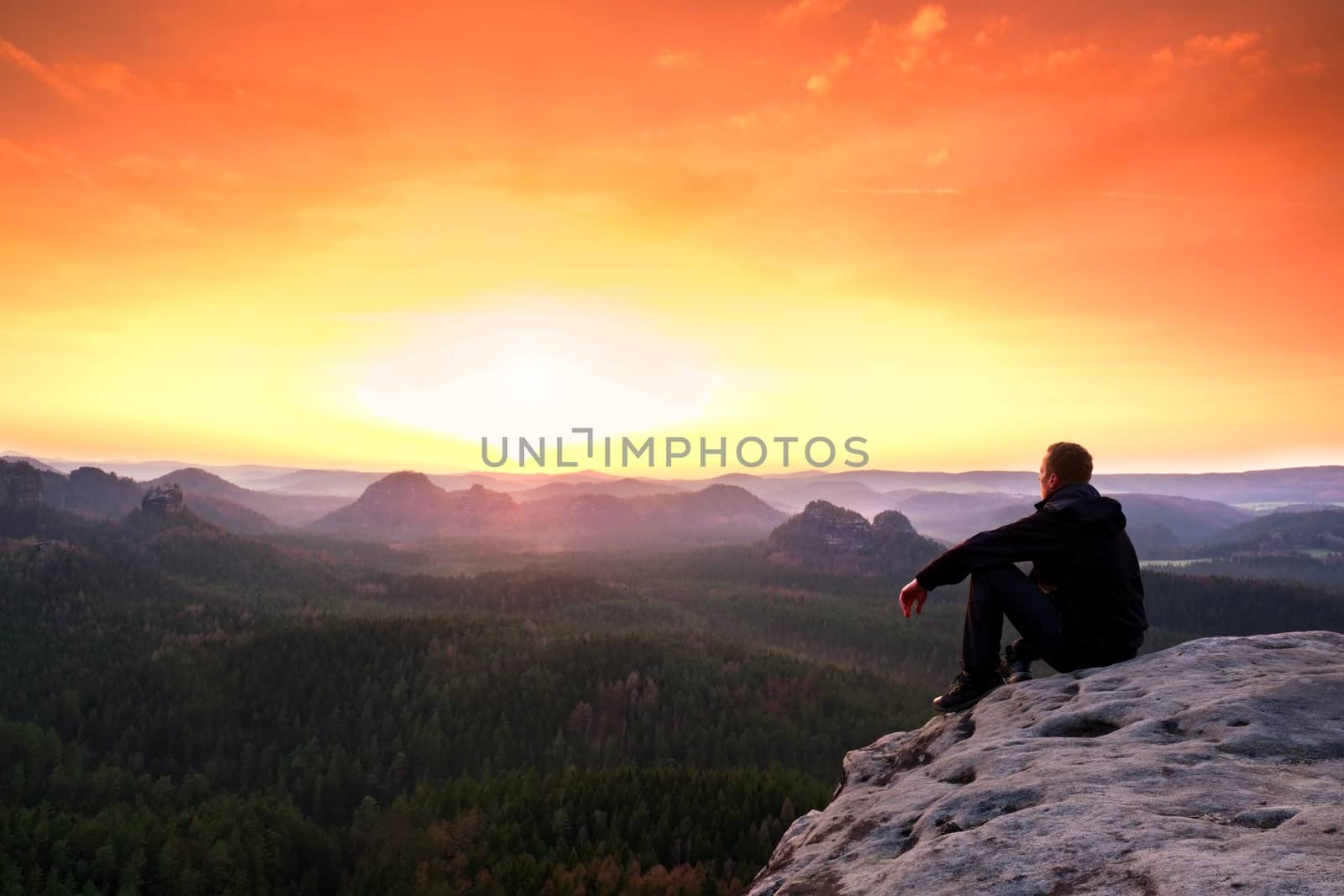 Adult tourist in black  sit on cliff's edge. Man enjoying morning  and looking to orange daybreak at misty horizon