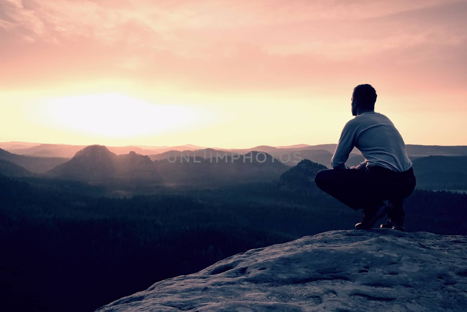 Adult hiker in black sit on mountain  edge. Man enjoying evening and looking to orange misty horizon
