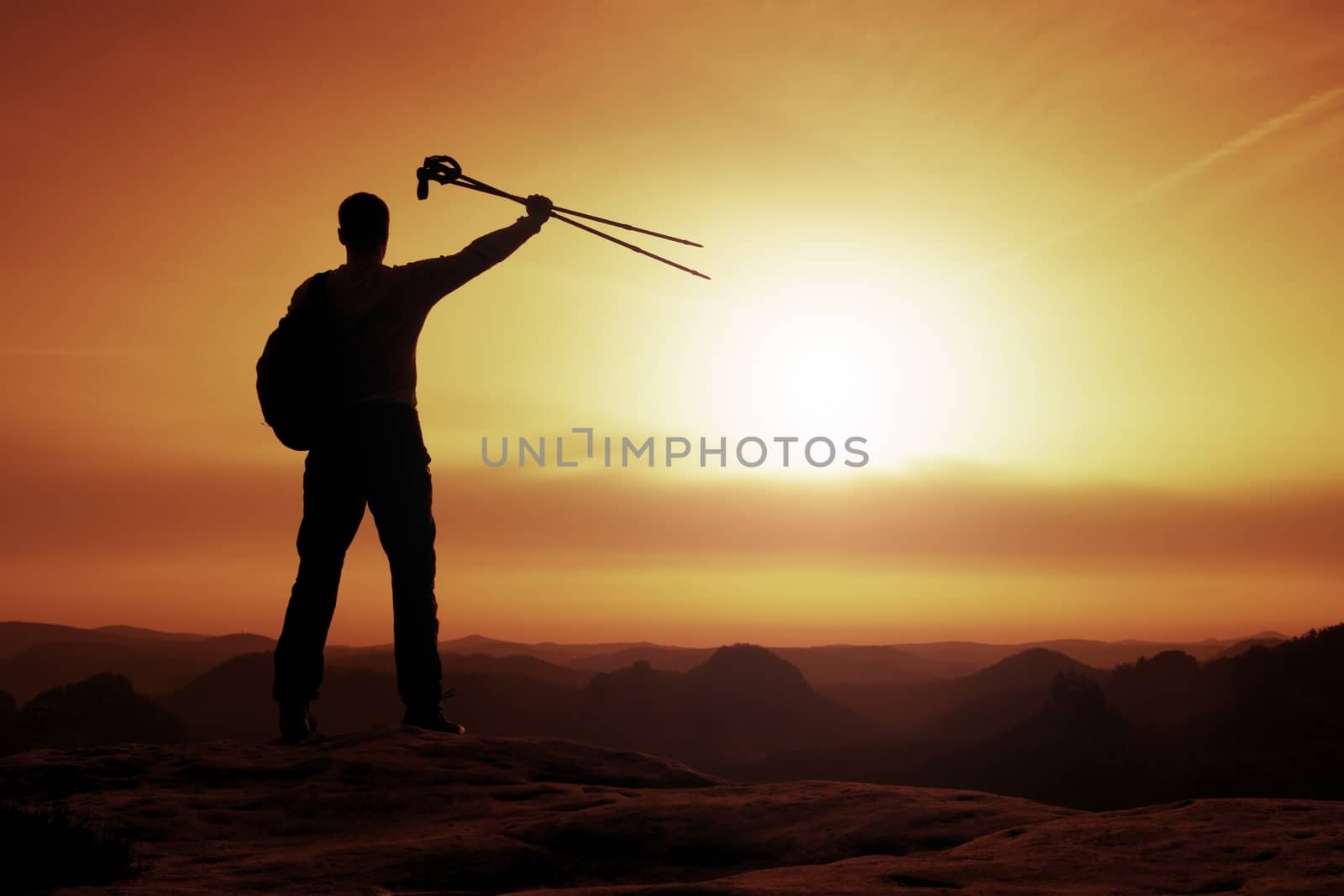 Silhouette of happy tourist with poles in hand above head. Sunny daybreak in mountains. Hiker with sporty backpack stand on rocky view point above misty valley. 