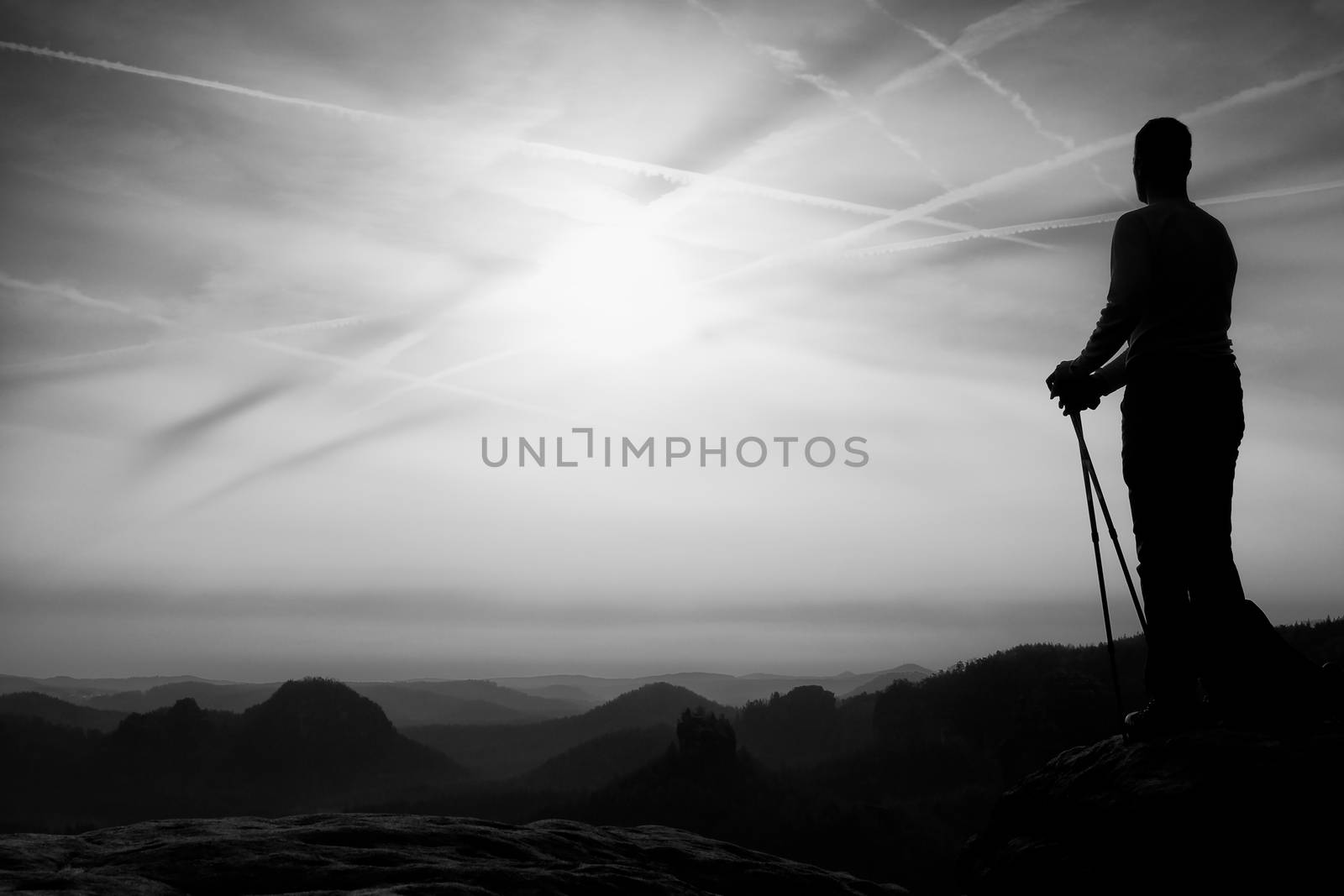 Silhouette of tourist with poles in hand. Sunny spring daybreak in rocky mountains. Hiker w on rocky view point above misty valley. 