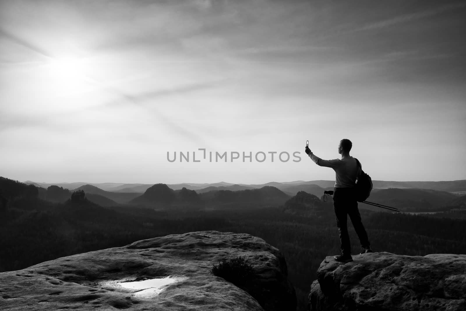 Tall tourist at dangerous cliff edge is taking selfie on peak above valley. Mountain peak and deep valley with sun in the frame