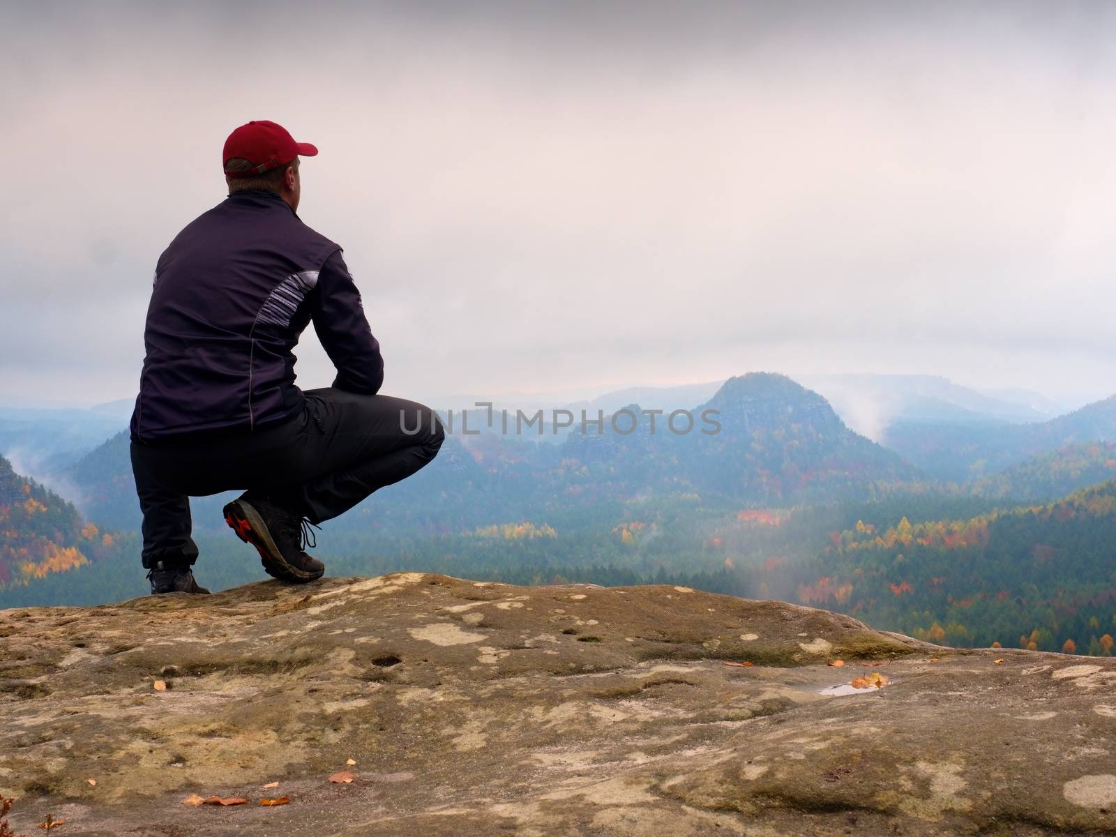 Young man in black sportswear is sitting on cliff's edge and looking to misty valley bellow. Autumn cold weather