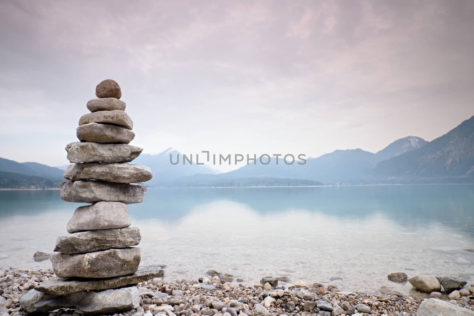 Balanced stone pyramide on shore of blue water of mountain lake. Blue mountains in water level mirror. Children built pyramid from pebbles.  Poor lighting conditions.