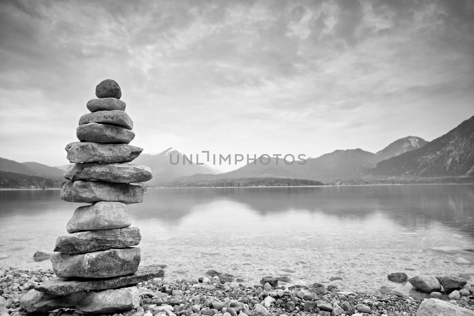 Balanced stone pyramide on shore of blue water of mountain lake. Blue mountains in water level mirror. Children built pyramid from pebbles.  Poor lighting conditions.