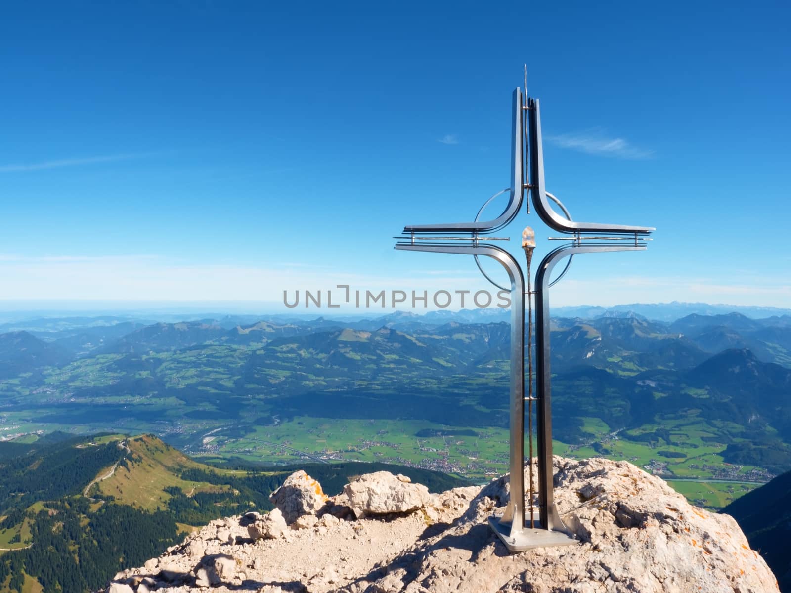Praying cross raised at mountain summit in Alps. Sharp peak, daybreak Sun in sky. Steel crucifix in memory of victims of mountains. Vivid photo.