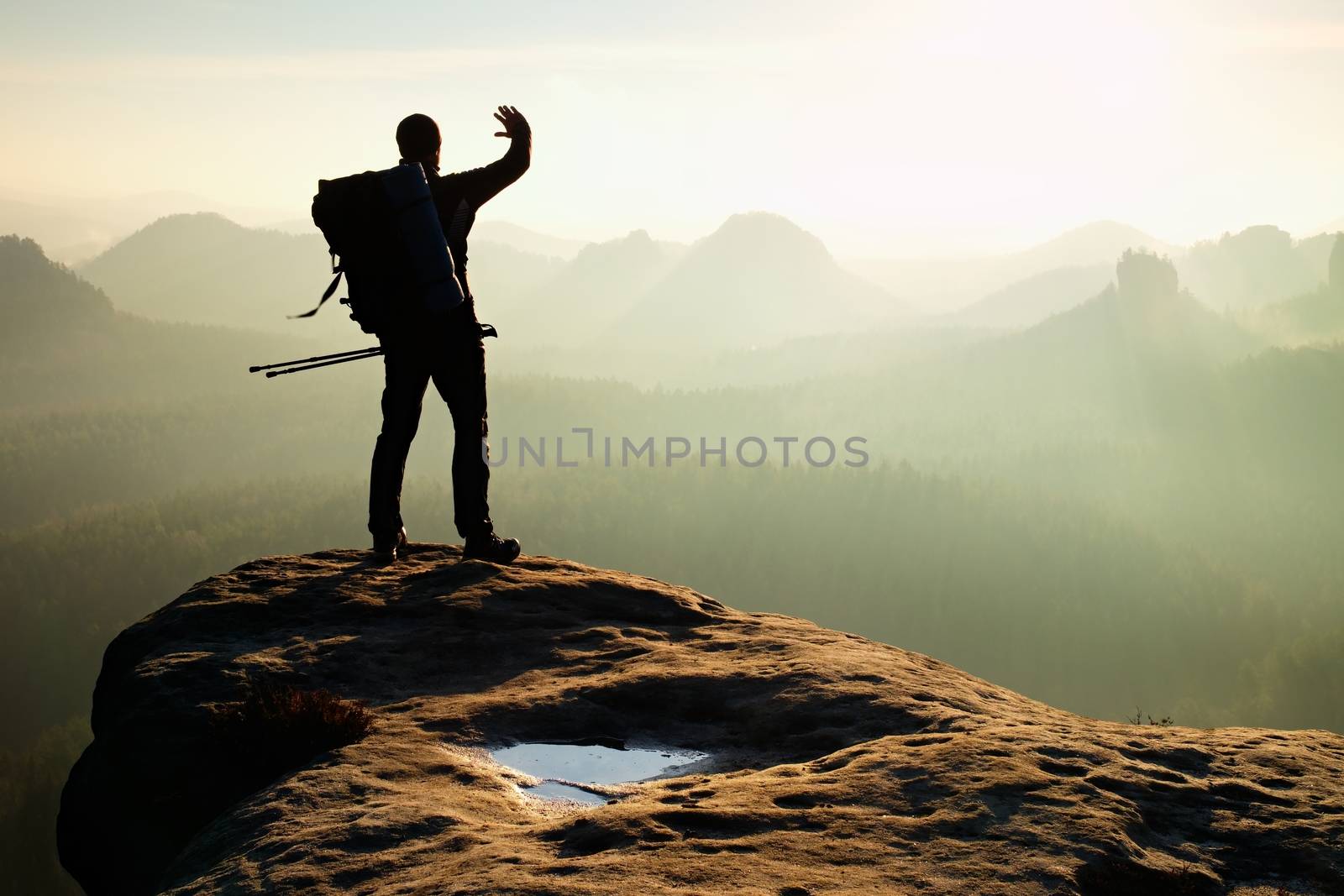 Hiker on peak. Backpacker with poles in hand shadowing eyes. Sunny spring daybreak in rocky mountains. Hiker on rocky view point above misty valley. 