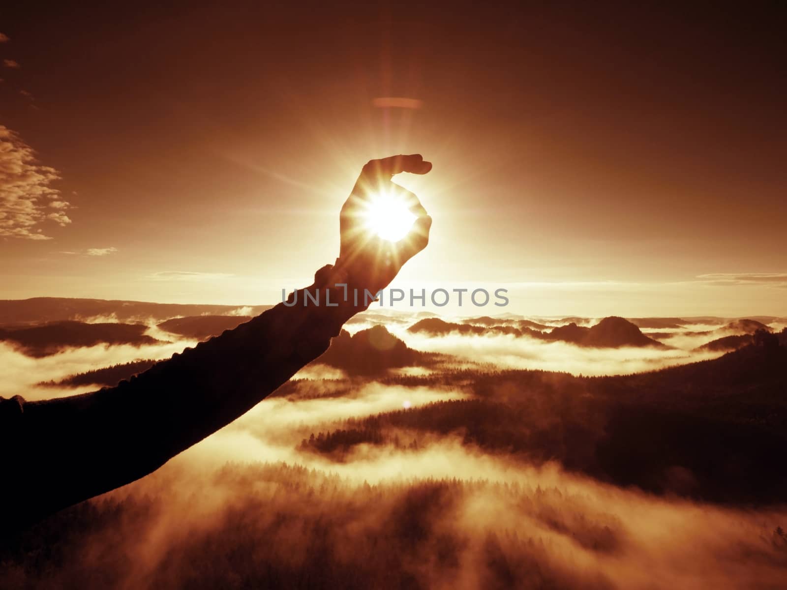 Man hand touch Sun. Misty daybreak in a beautiful hills. Peaks of hills are sticking out from foggy background, the fog is red and orange due to Sun rays. 