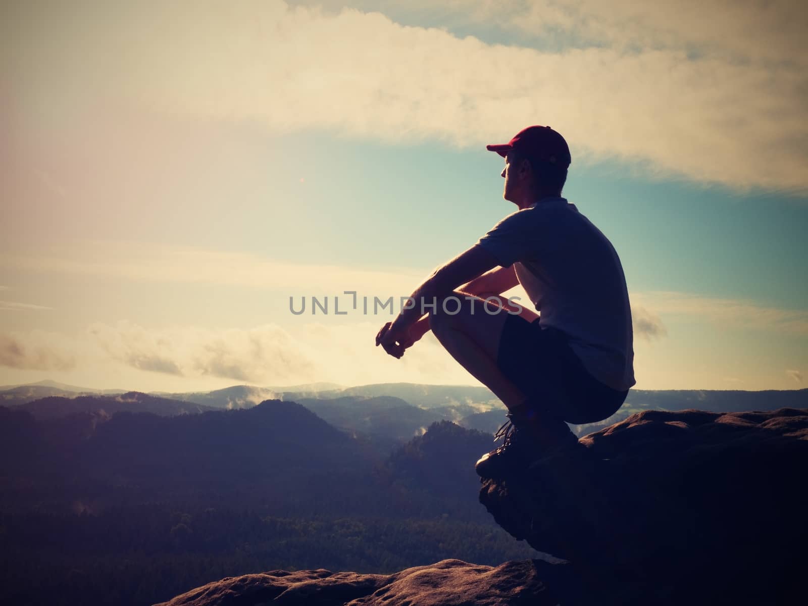 Hiker man take a rest on mountain peak. Man sit on summit, bellow spring valley. Bright morning Sun shining in sky. 