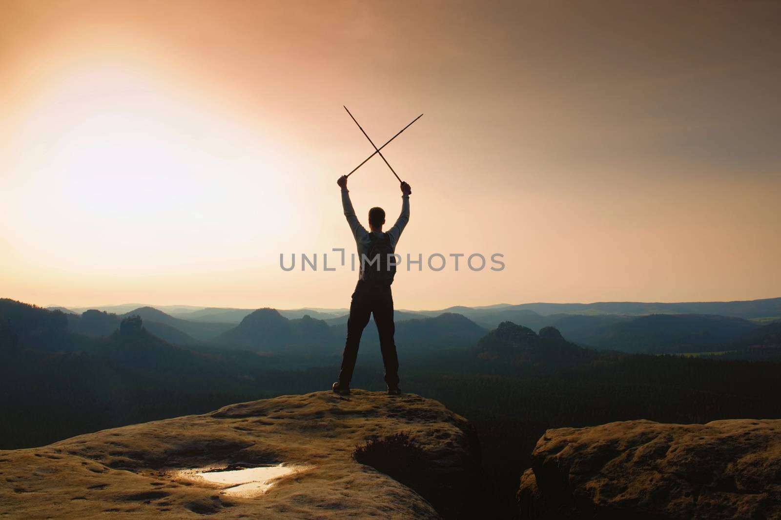 Man with crossed poles above head on cliff.  Foggy  mountain valley bellow. Happy tourist guide with poles in hands. Sunny spring daybreak in hills.