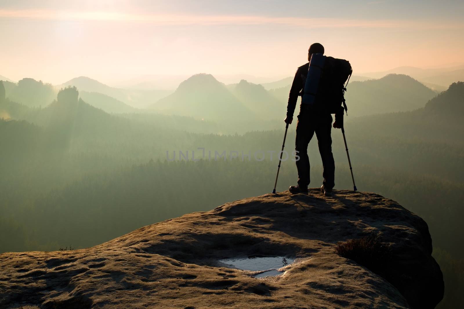 Tourist with leg in immobilizer. Hiker silhouette with medicine crutch on mountain peak. Deep misty valley bellow silhouette of man with hand in air. Spring daybreak