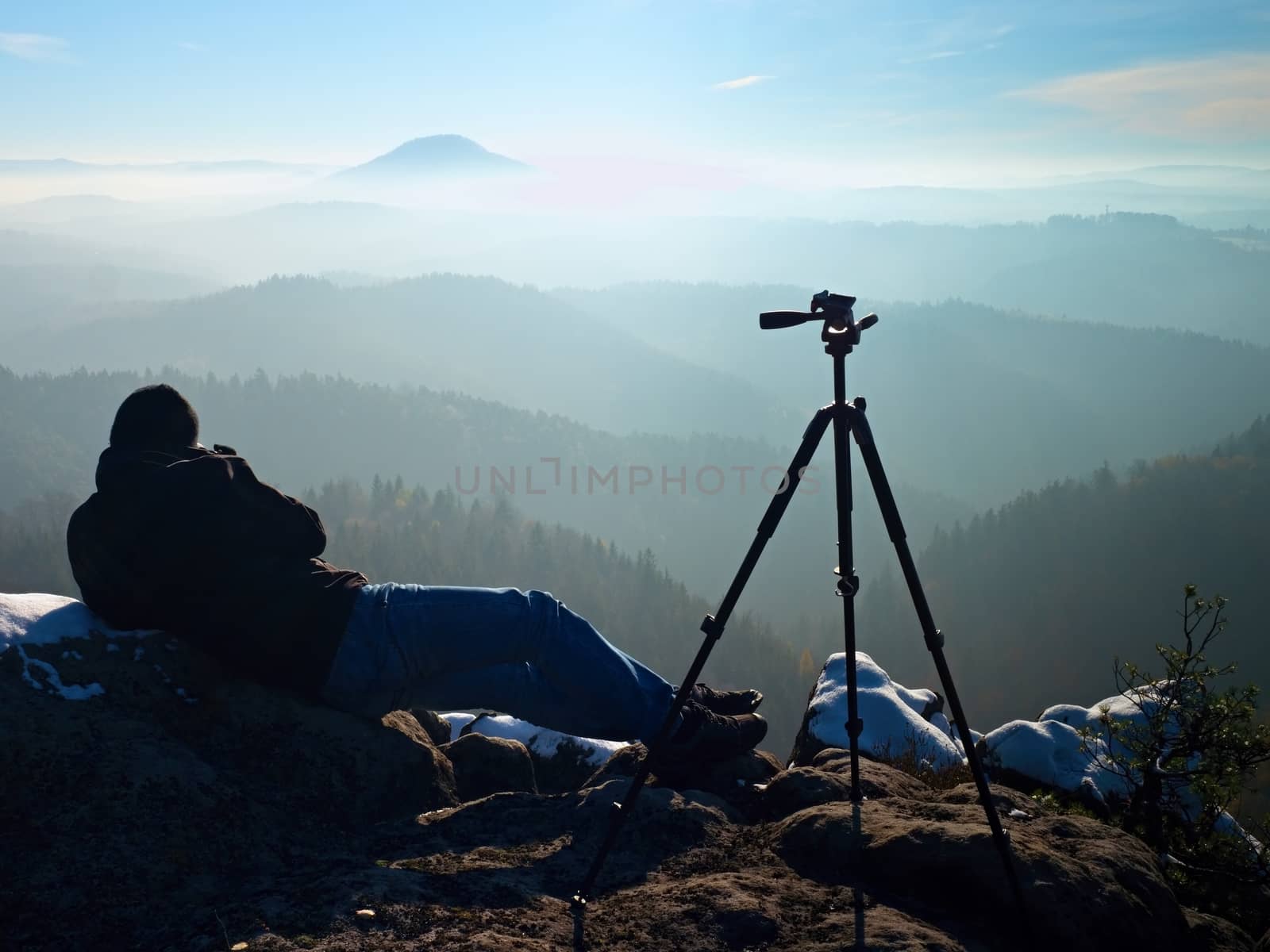 Professional nature photographer do work on snowy cliff.  Man takes photos with mirror camera on peak of rock. Dreamy fogy landscape, spring orange pink misty sunrise.