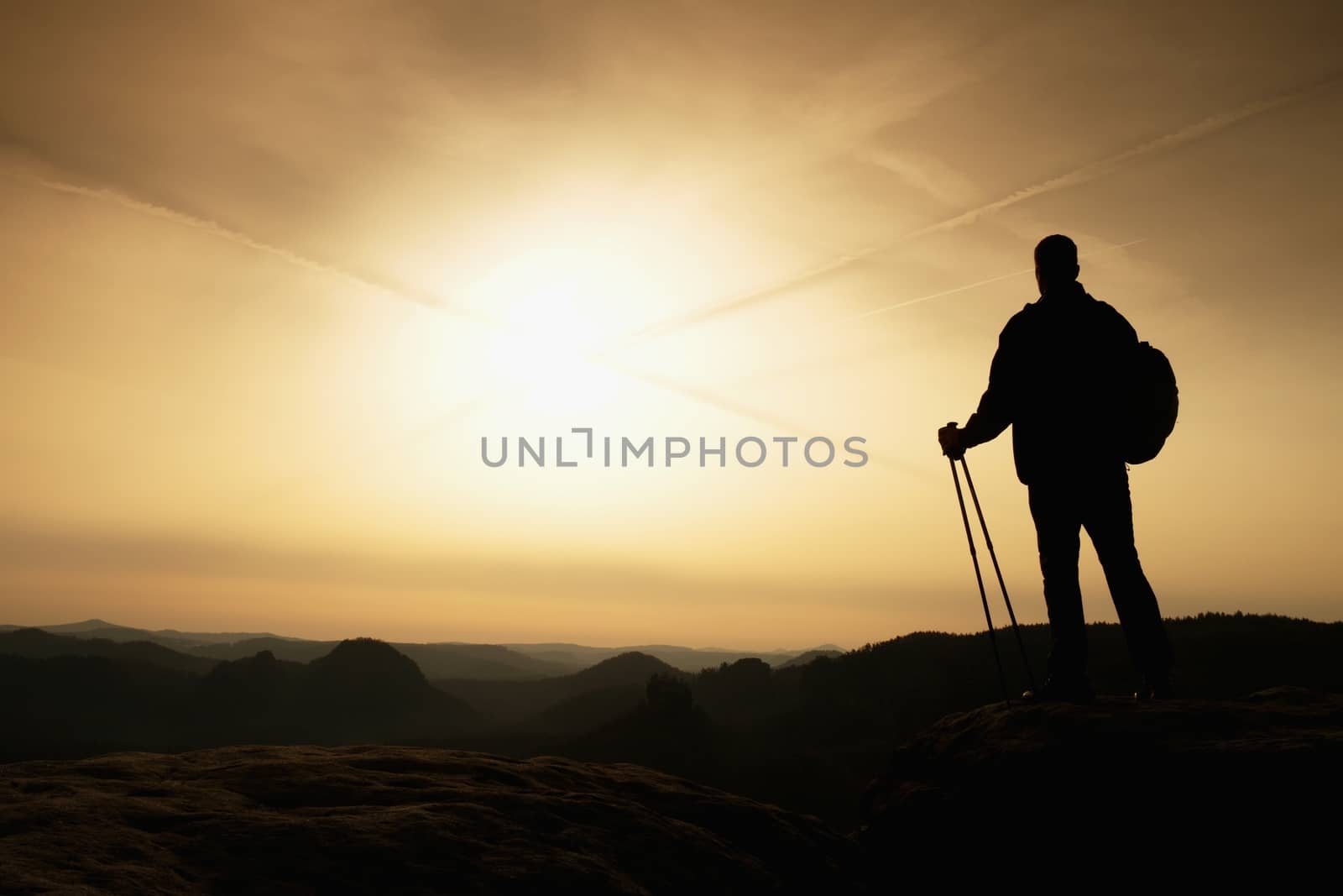 Tall backpacker with poles in hand. Sunny spring daybreak in rocky mountains. Hiker with big backpack stand on rocky view point above misty valley. 