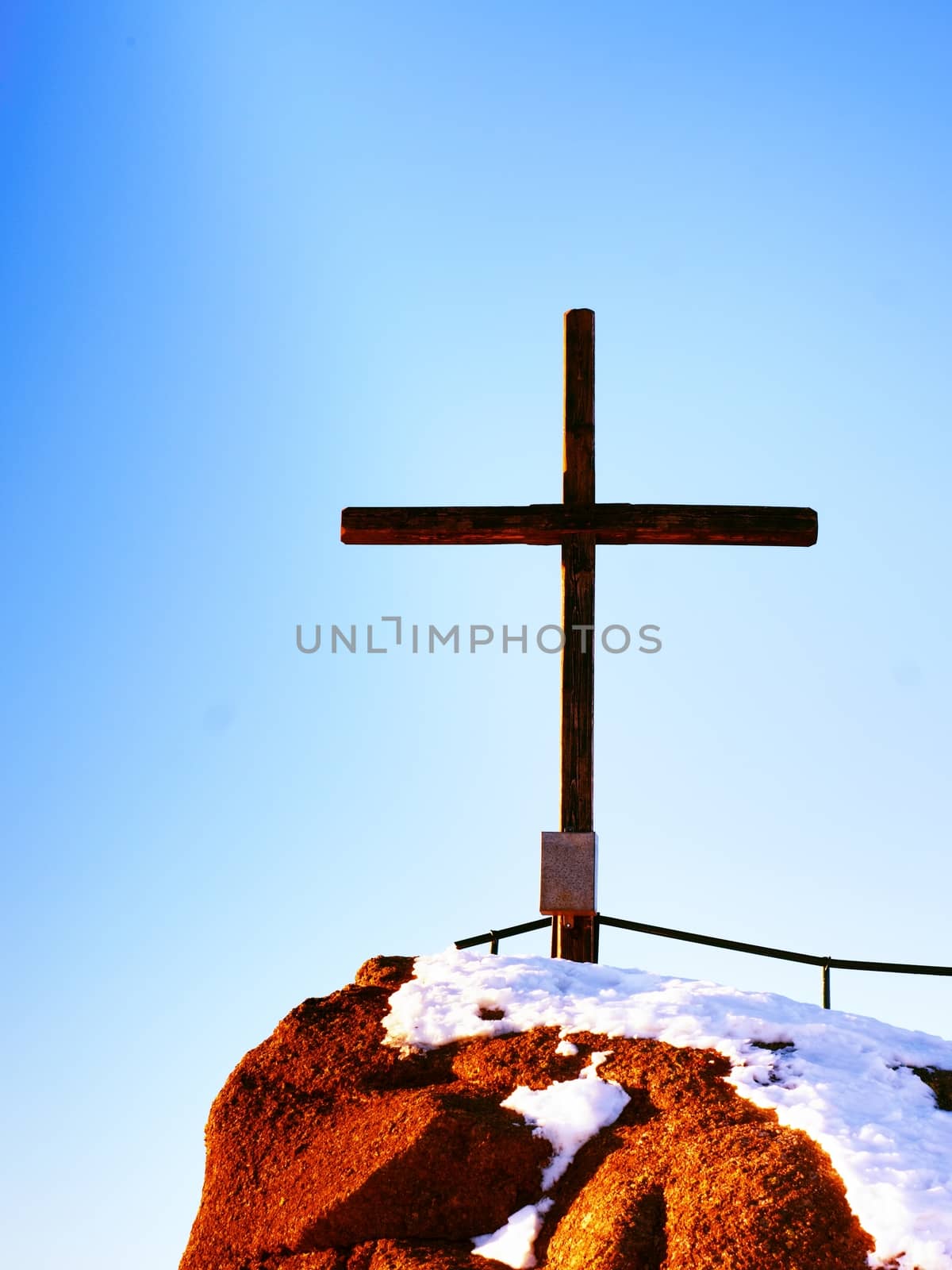 Modest wooden cross raised  on rocky mountain summit . Sharp snowy peak. Daybreak Sun in sky. Wooden unpretentious crucifix in memory of victims of mountains. Vivid photo.