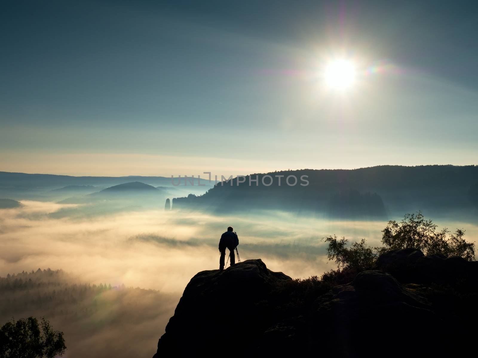 Nature photographer with tripod and camera on cliff and thinking. Dreamy fogy landscape, orange misty sunrise in a beautiful mountains. Vivid and strong vignetting effect.